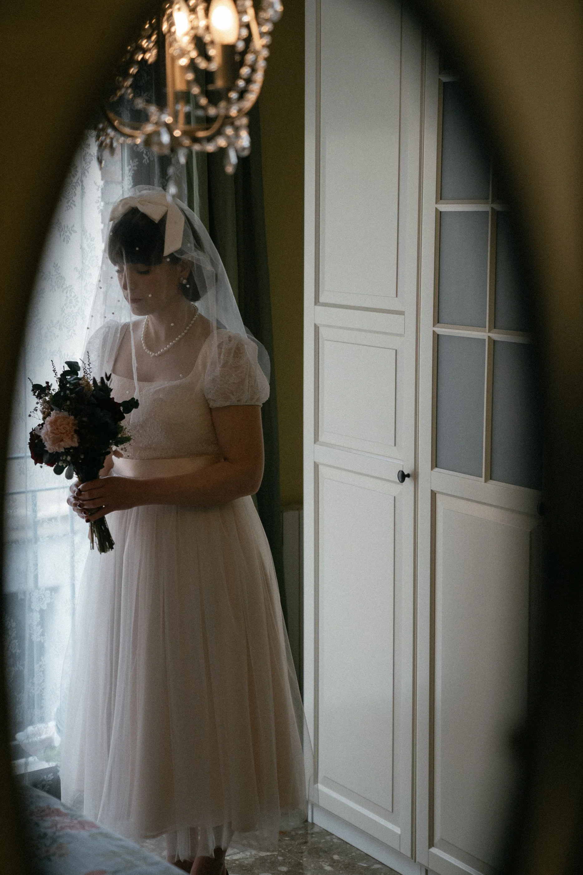 A bride wearing a vintage-style white wedding dress, a pearl necklace, and a veil, holding a bouquet of dark and light flowers, standing in a room seen through a mirror.