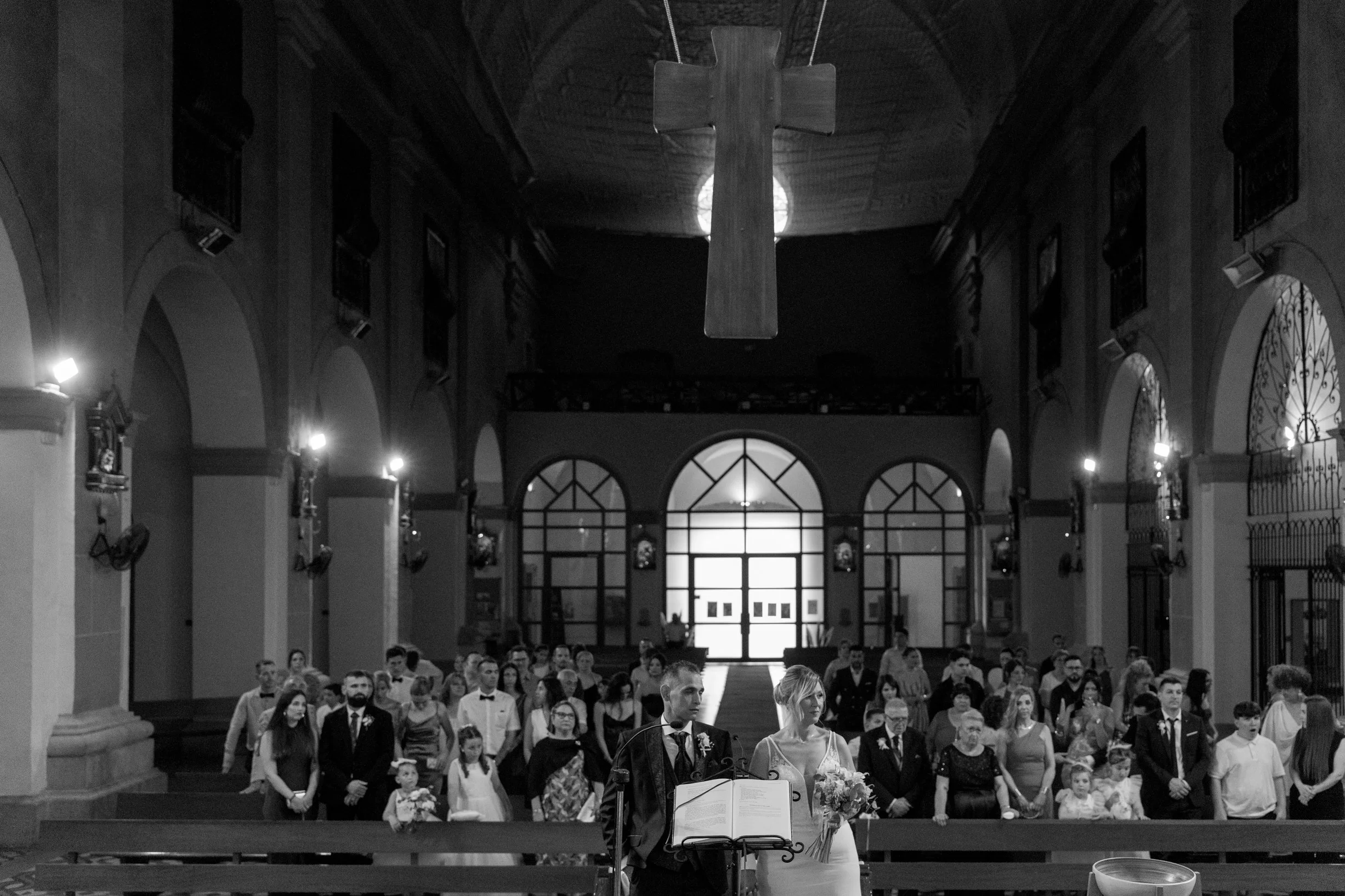 A black and white photo of a wedding ceremony inside a church. The bride and groom are standing at the altar, holding hands. Behind them, guests are seated, facing forward. The church has tall arches and large windows, with a high ceiling and a large