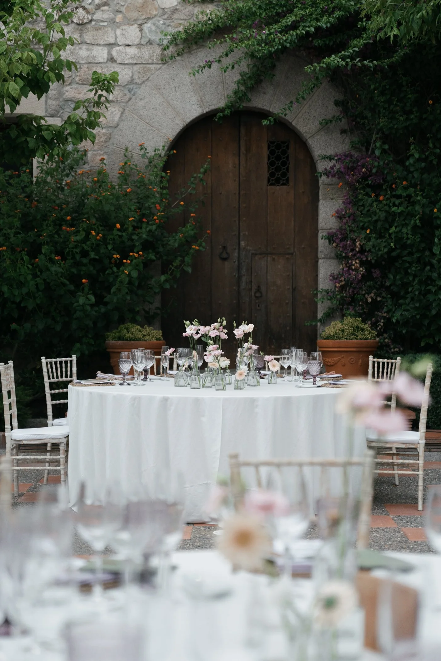 Elegant outdoor dining setup with a round table covered with a white tablecloth, decorated with pink flowers in glass vases, surrounded by white chairs, set against a rustic stone and wood door with greenery and flowering plants.