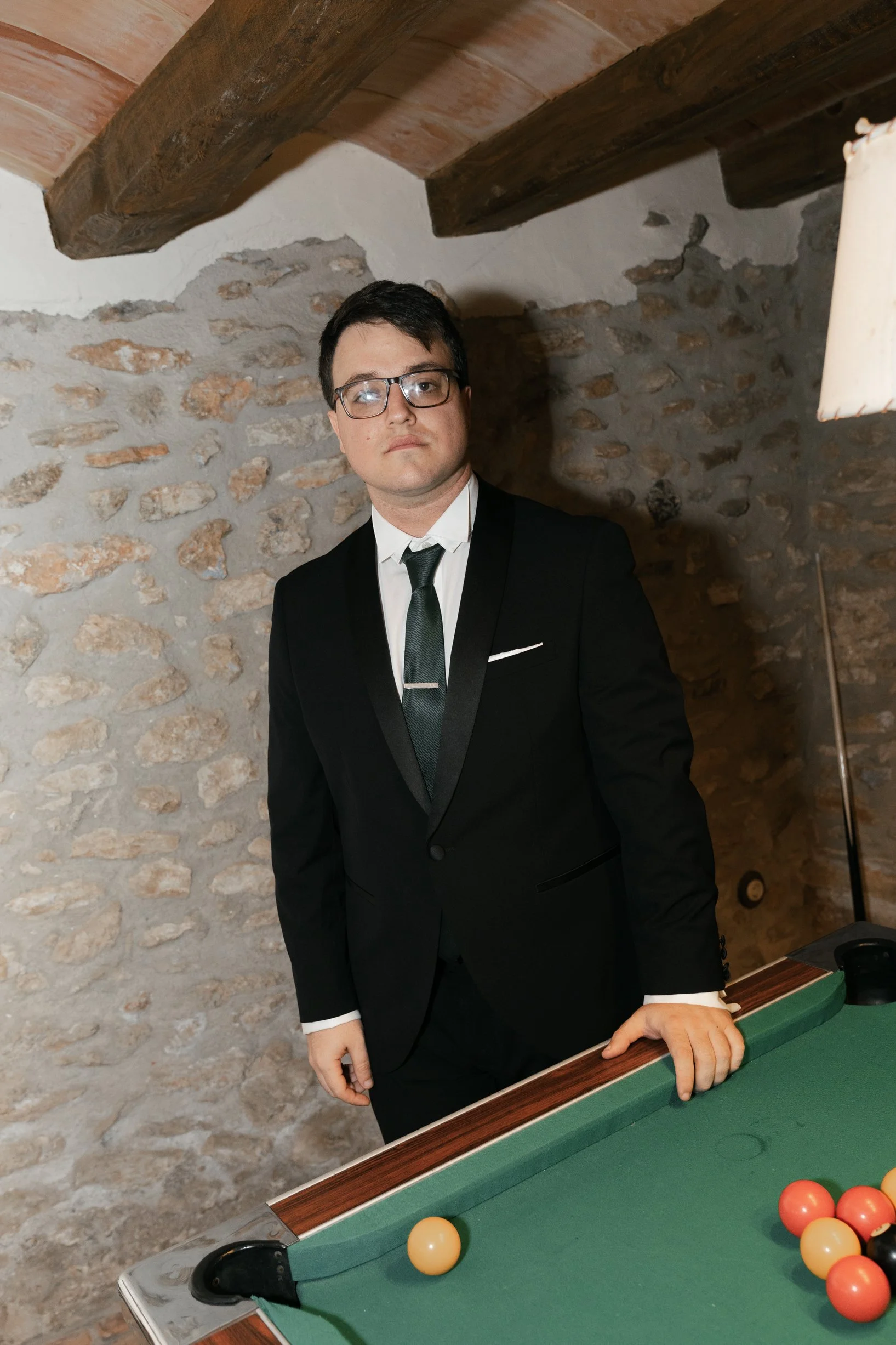A man in a black tuxedo and glasses standing next to a pool table in a room with stone walls and wooden beams.
