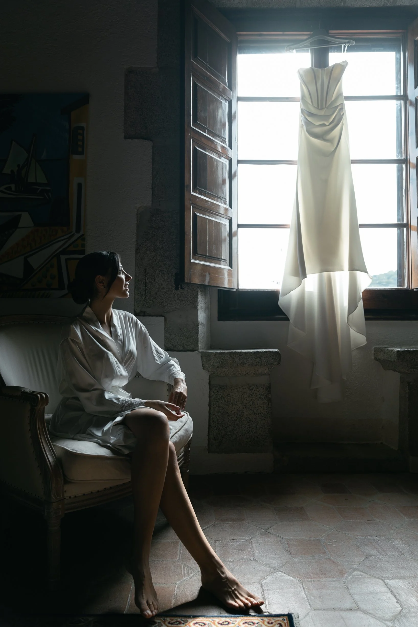A woman sitting on a vintage-style sofa, looking at a wedding dress hanging in front of a window with open wooden shutters, backlit by daylight.