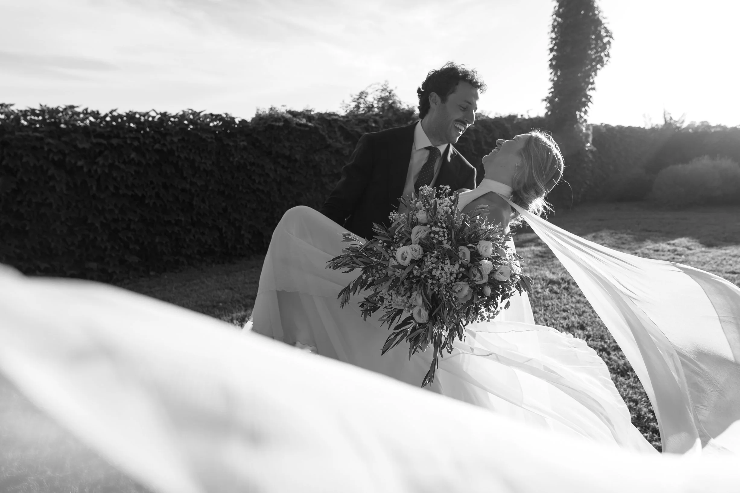 A newlywed couple enjoying a moment outdoors, with the groom holding the bride who is holding a bouquet of flowers, both smiling and looking at each other, in black and white.