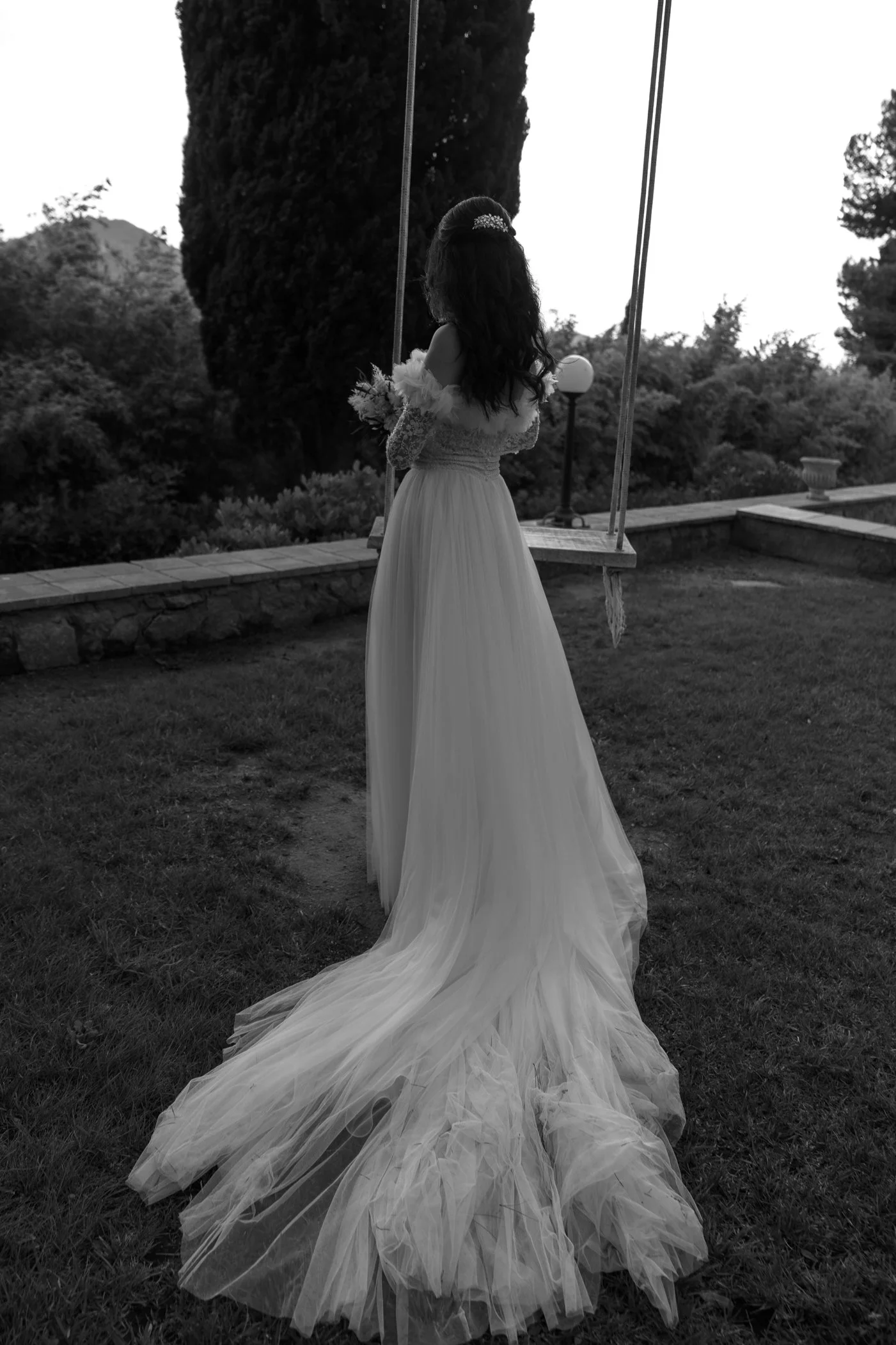 A woman in a wedding dress standing on grass by a swing, holding a bouquet, with trees and a stone wall in the background.