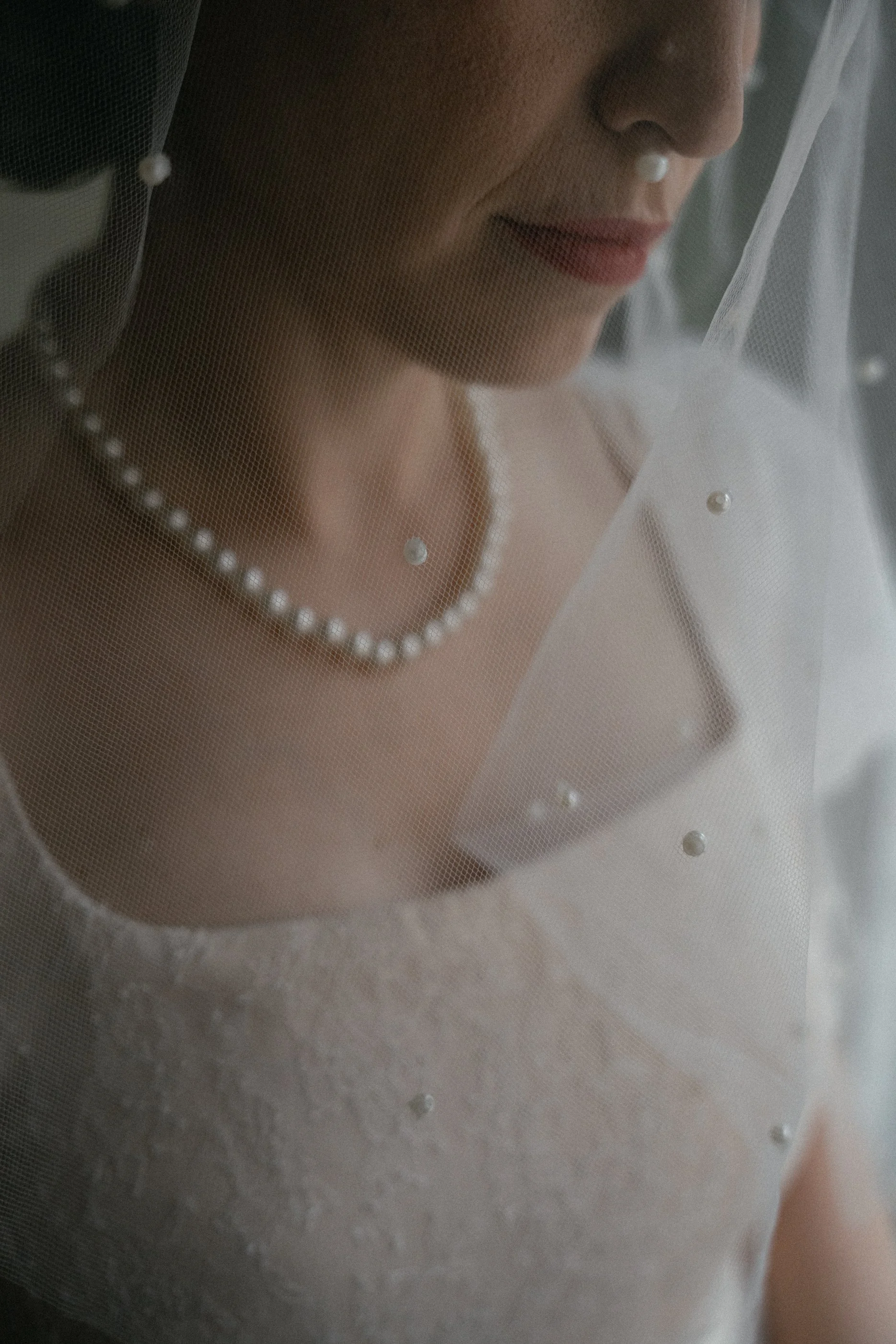Close-up of a woman's face and upper torso, wearing a pearl necklace and a white garment, behind a sheer fabric decorated with small pearls, creating a soft, elegant portrait.