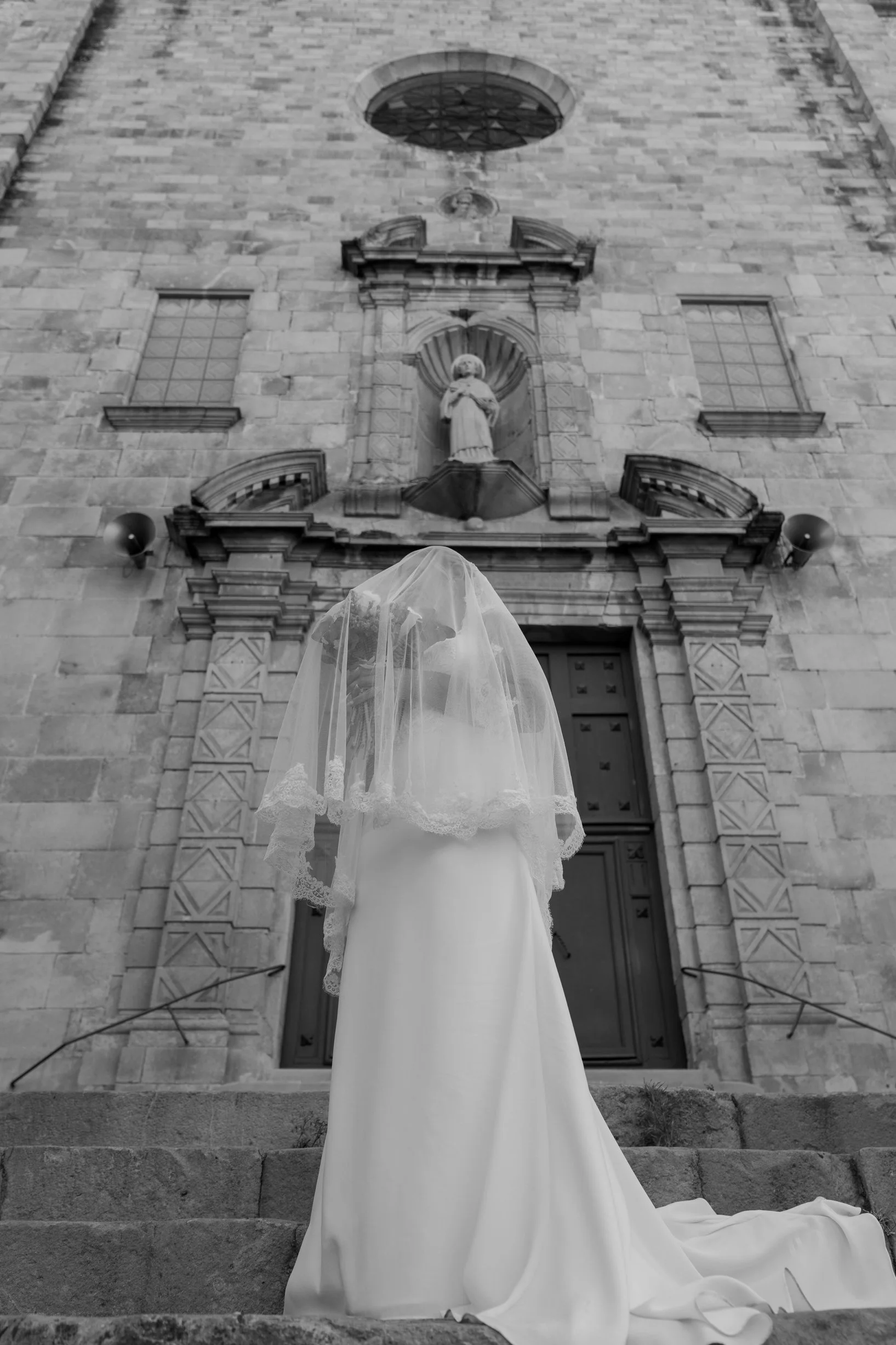 A bride in a wedding dress and veil stands on stairs outside a historic stone church, facing the building.