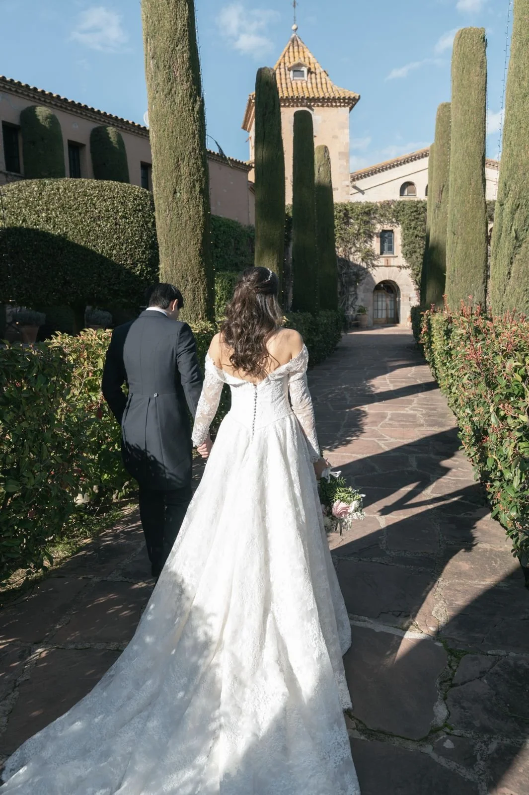 A bride and groom walking hand in hand on a sunny pathway with tall cypress trees and a historic building in the background.