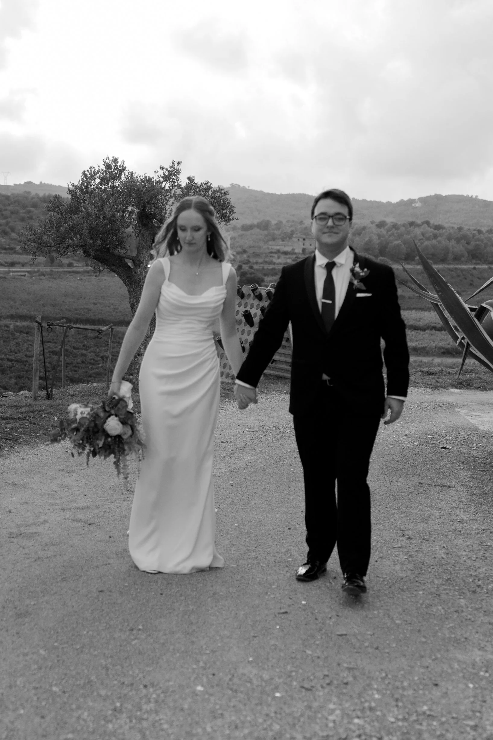 Black and white photo of a bride and groom holding hands outdoors, walking on a dirt path, with trees and hills in the background.