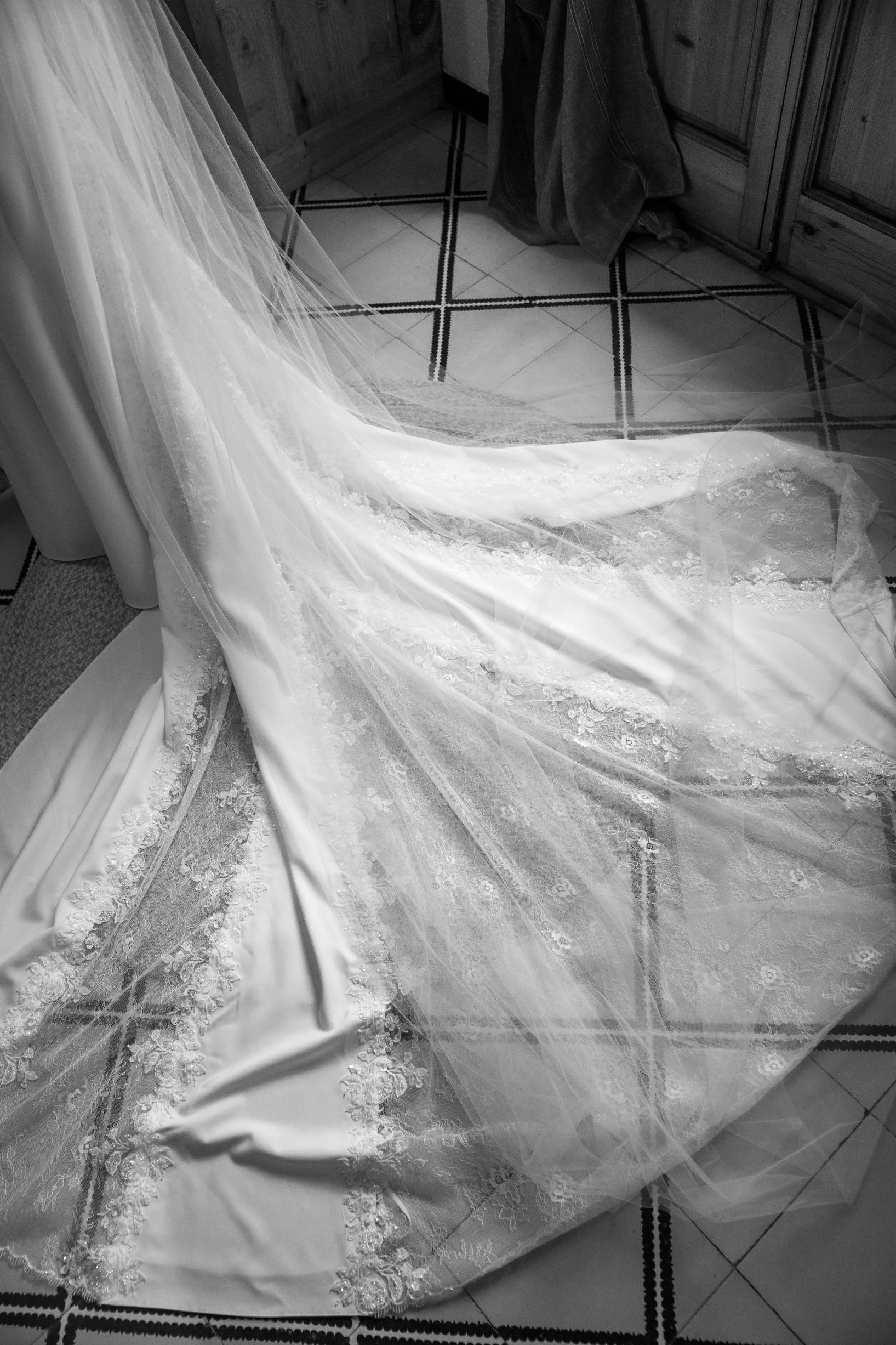 A wedding dress with intricate lace and embroidery on a tiled floor near a wooden cabinet.