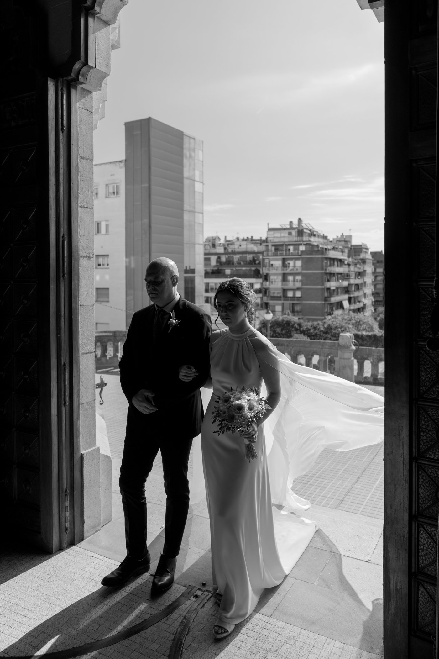 Black and white photo of a bride in a wedding dress holding a bouquet, walking with a man, likely her father, through an open doorway with city buildings in the background.