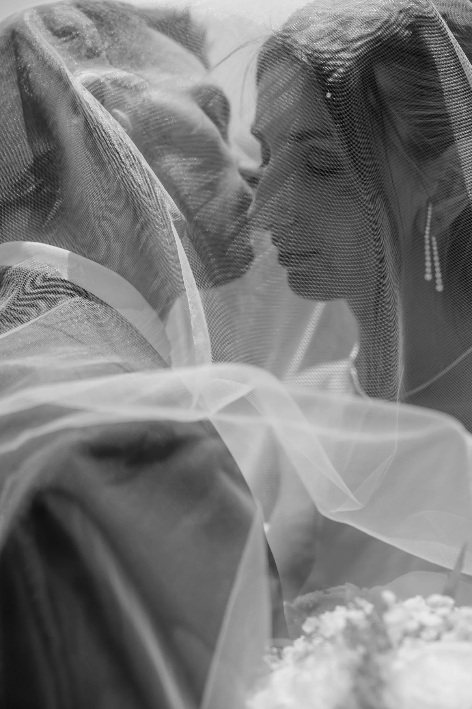 A woman and a man face-to-face, close together, with their eyes closed, partially covered by sheer fabric, in black and white photography.