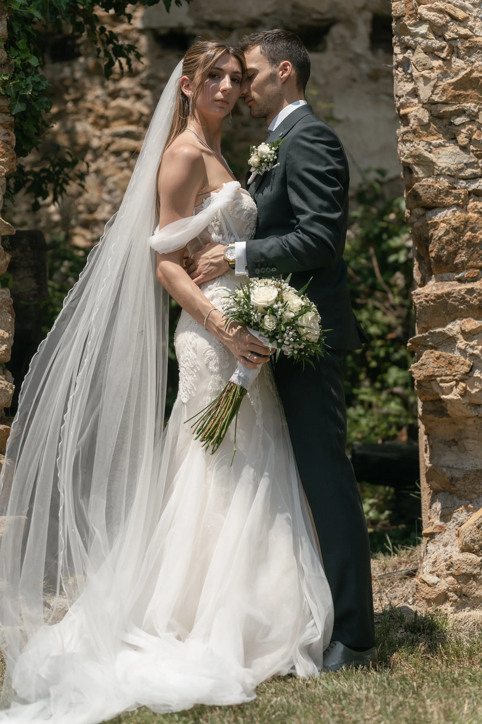 A bride and groom in wedding attire standing close, with the bride holding a white bouquet, outdoors among stone ruins, sharing an intimate moment.
