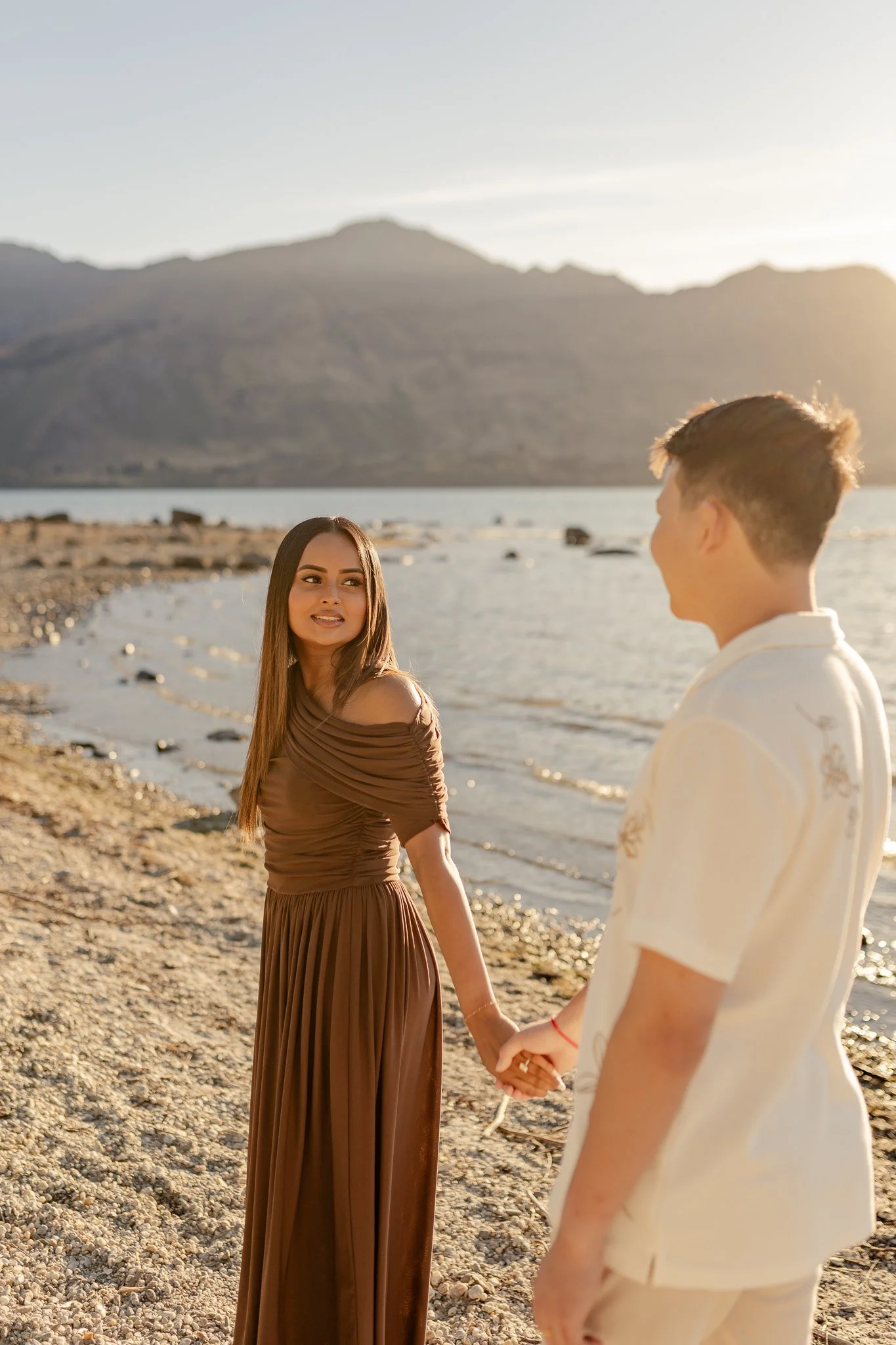 A young woman in a brown dress and a young man in a cream shirt hold hands while standing on a rocky beach at sunset, with mountains and water in the background.