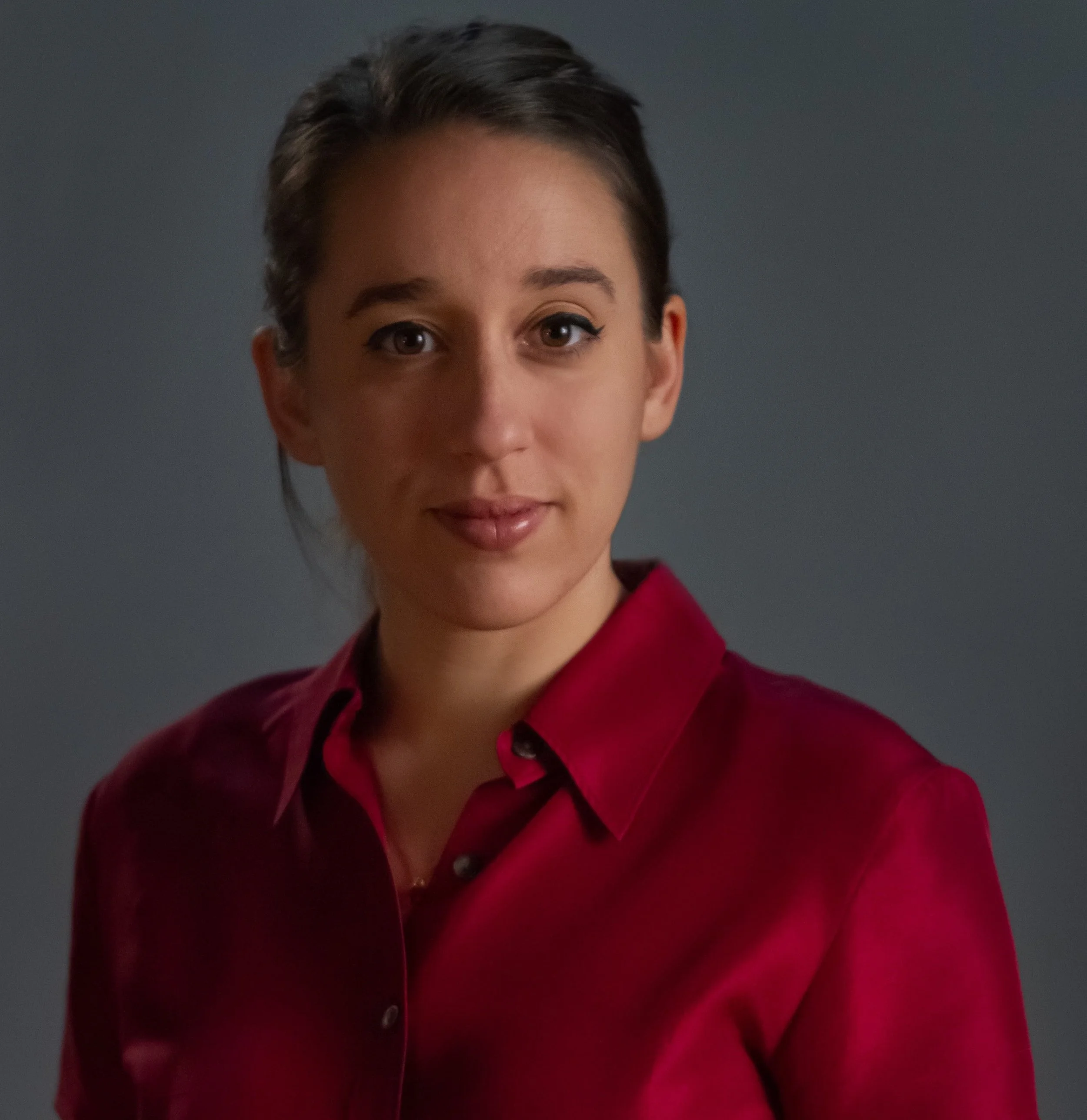 A white woman with brown hair tied back. She is wearing a red shirt and smiling directly to camera.