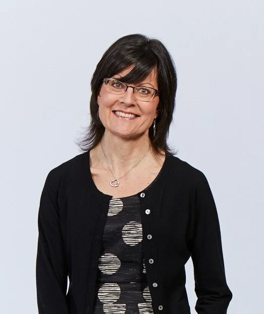 A white woman with shoulder length brown hair. She is wearing glasses, silver earrings and necklace, a black top with large white spots and a black cardigan. She is smiling to the camera.