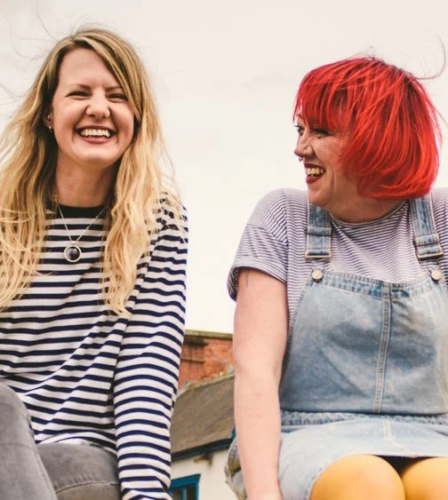 Two white women, one with long blonde hair, another with bright, red hair, are sat side by side laughing.