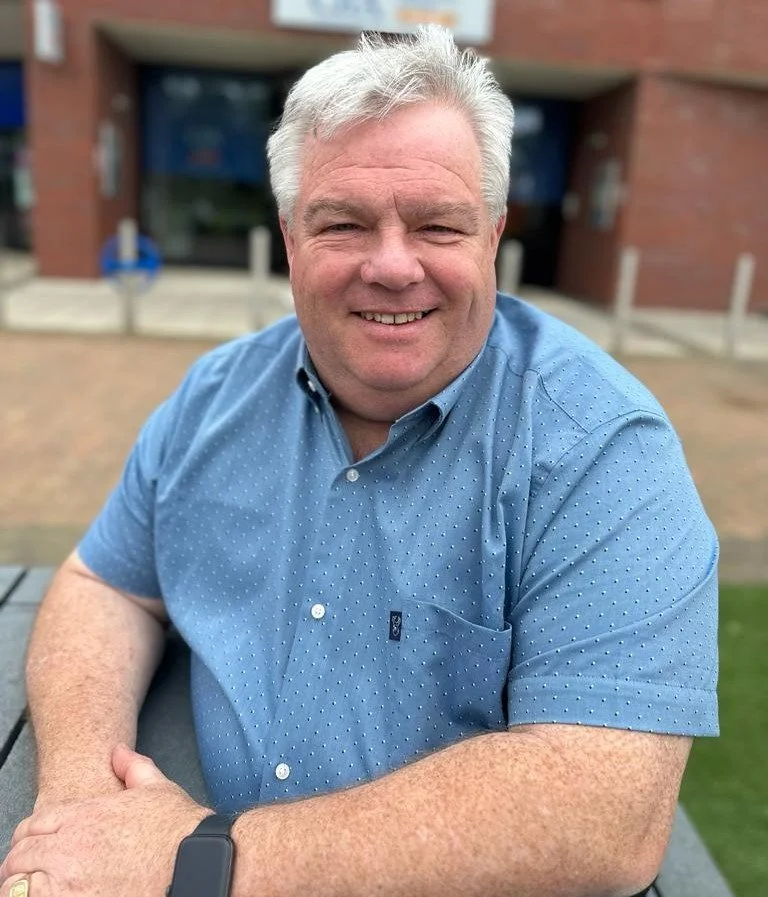 White man with grey hair looking to camera and smiling. He is wearing a blue short sleeve shirt and a black watch.