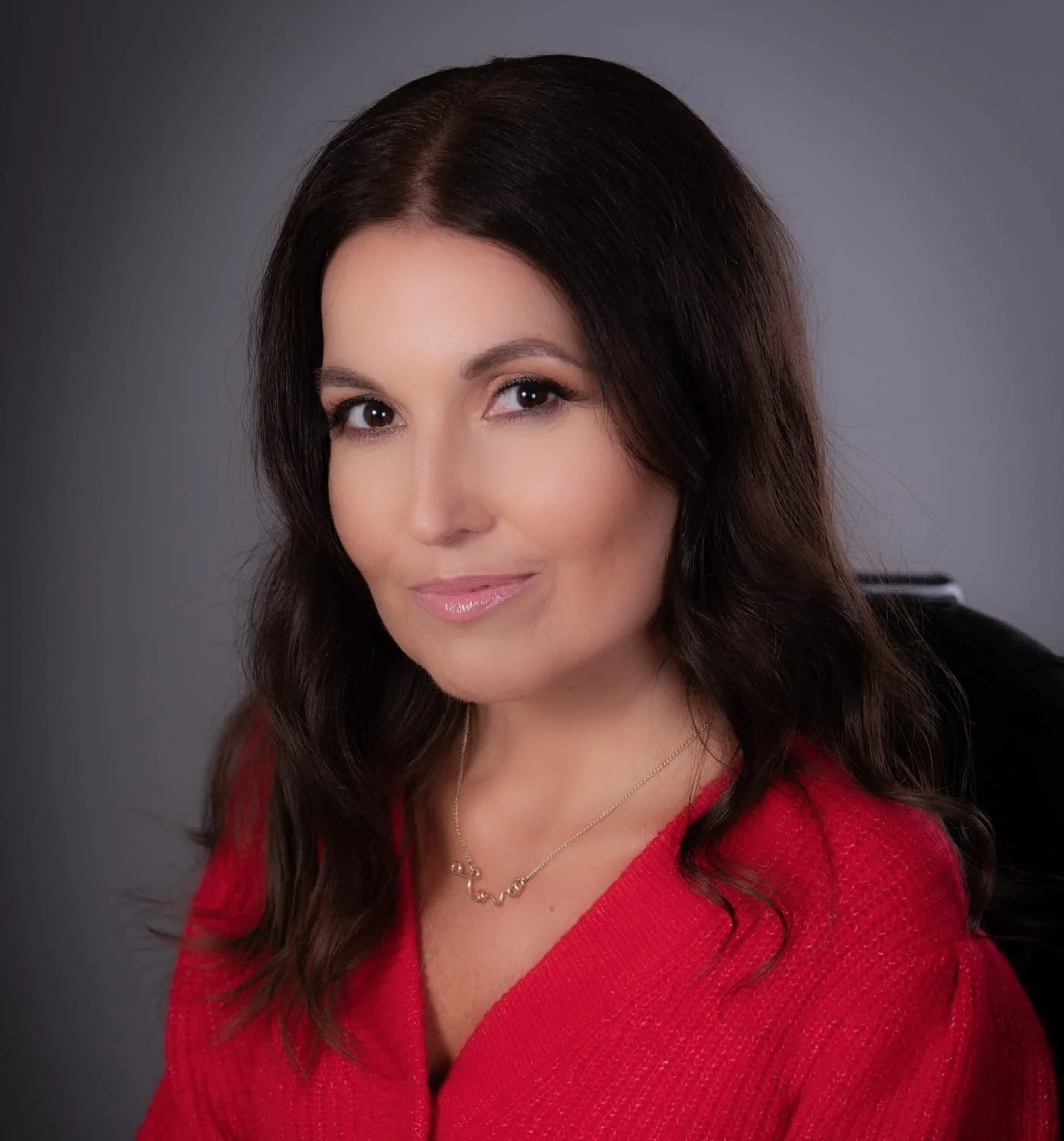 White woman with long brown wavy hair, smiling to camera. She is wearing a red jumper and a gold necklace.