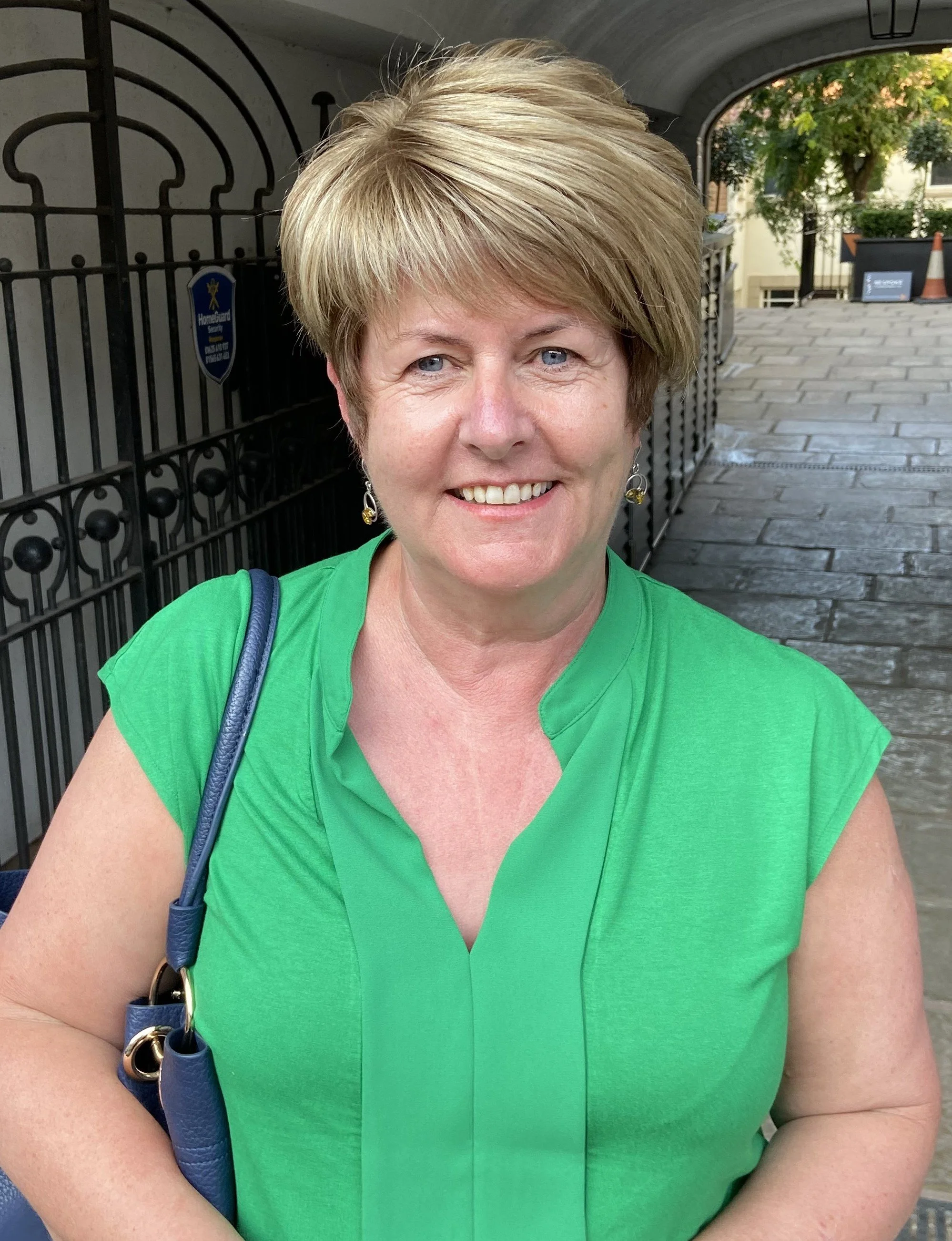 White woman with blonde, short hair, blue eyes, smiling to camera. She is wearing a green blouse, gold earrings and is carrying a blue bag on her shoulder.