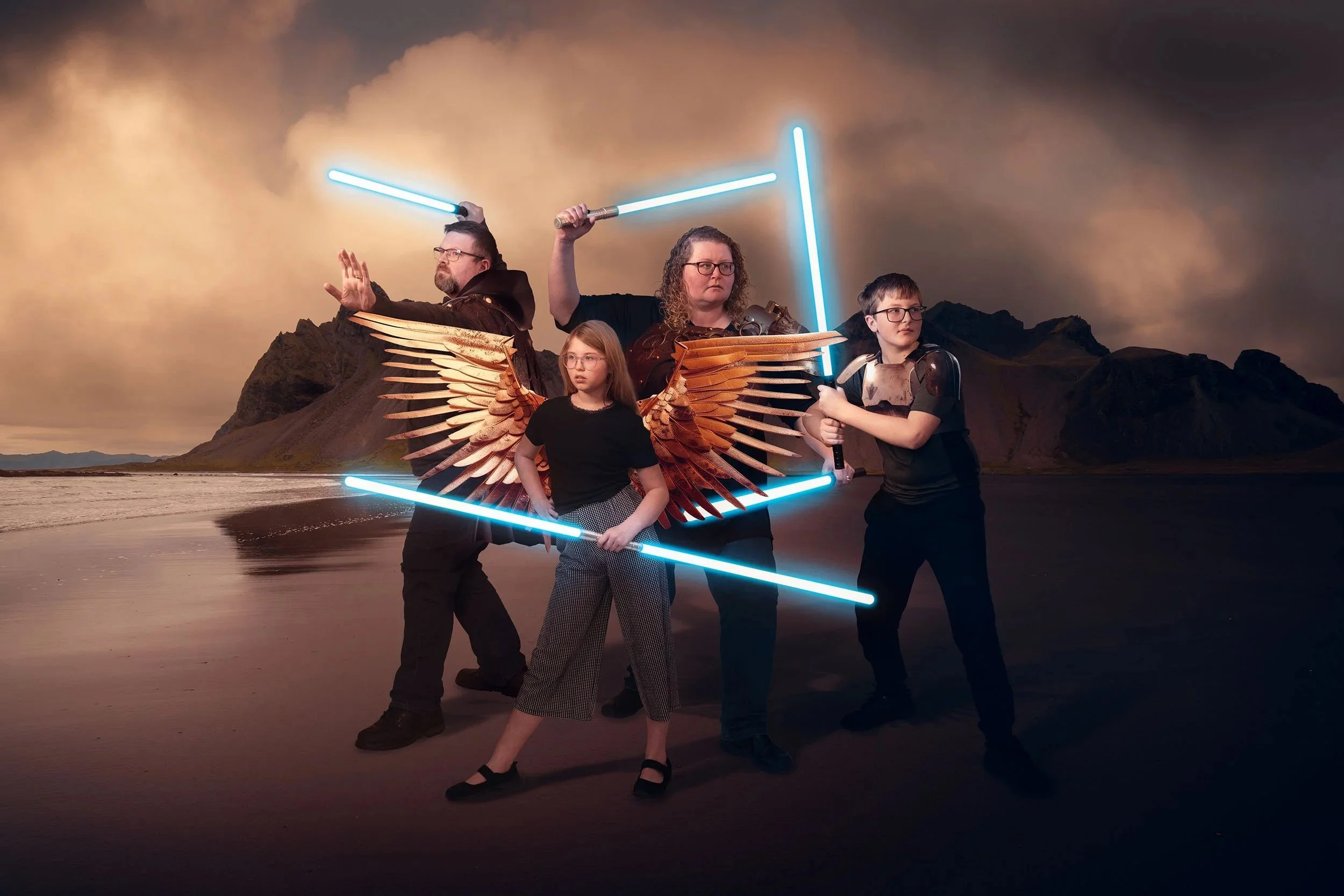 Four people dressed as sci-fi characters with glowing lightsabers, angel wings, and futuristic armor, standing on a beach with mountains and an overcast sky in the background.