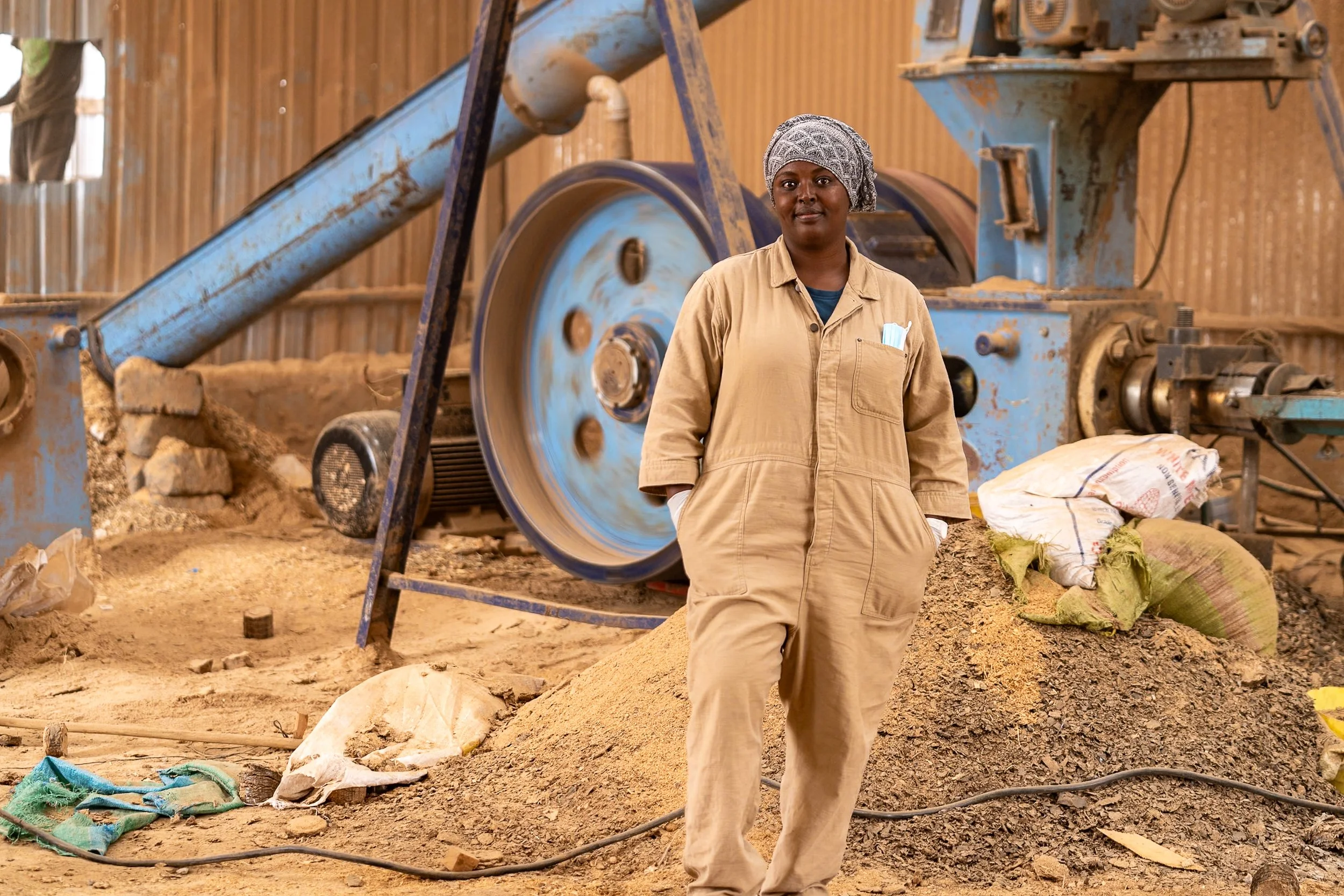 A woman in a beige jumpsuit and a patterned headscarf stands with her hands in her pockets in front of a large industrial machine and piles of sand, dirt, and construction materials inside a workshop.