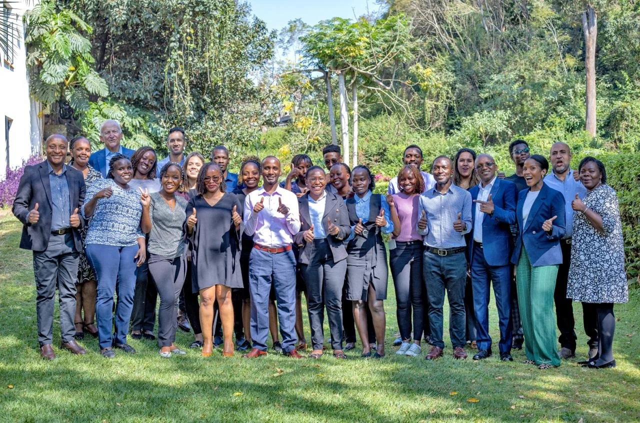 A diverse group of men and women dressed in business attire standing outdoors on grass, smiling and giving thumbs up, with trees and greenery in the background.