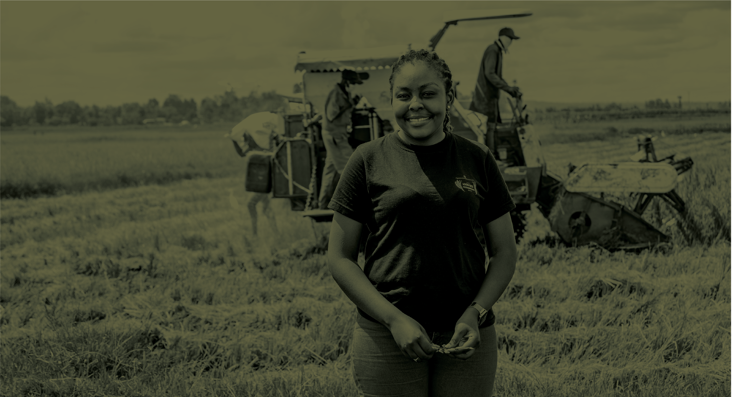 A young woman standing in a field of crops with a machine operating in the background.