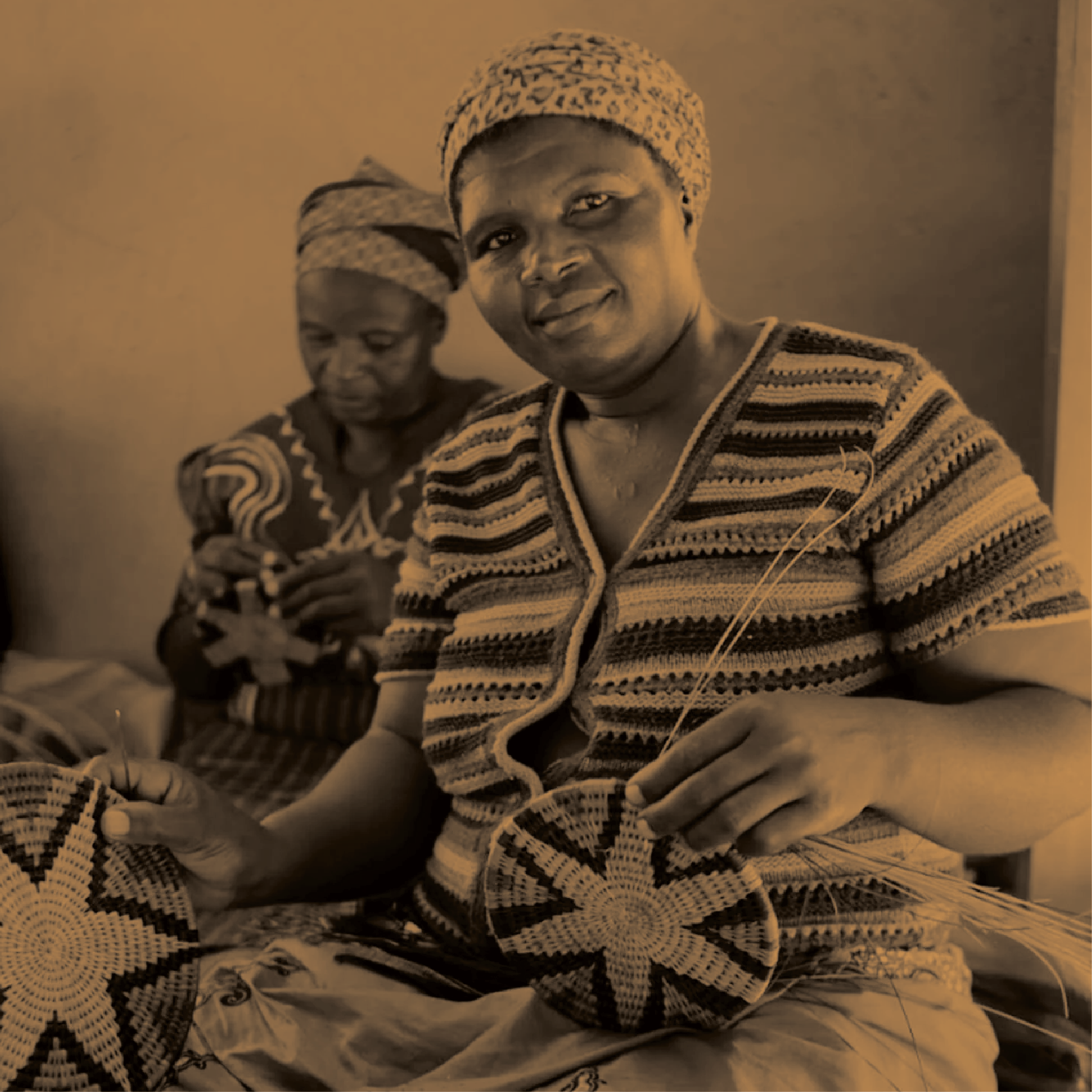 Two women, sitting together indoors, are engaged in traditional basket weaving, with one woman in the foreground smiling at the camera and holding a partially completed woven item, and the other woman in the background focused on her work.