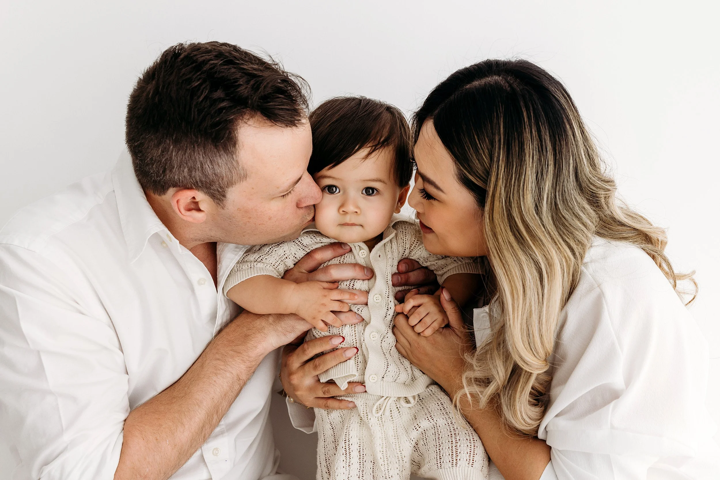 Mum and dad cuddling and kissing their baby during a family session in the Aveley studio.