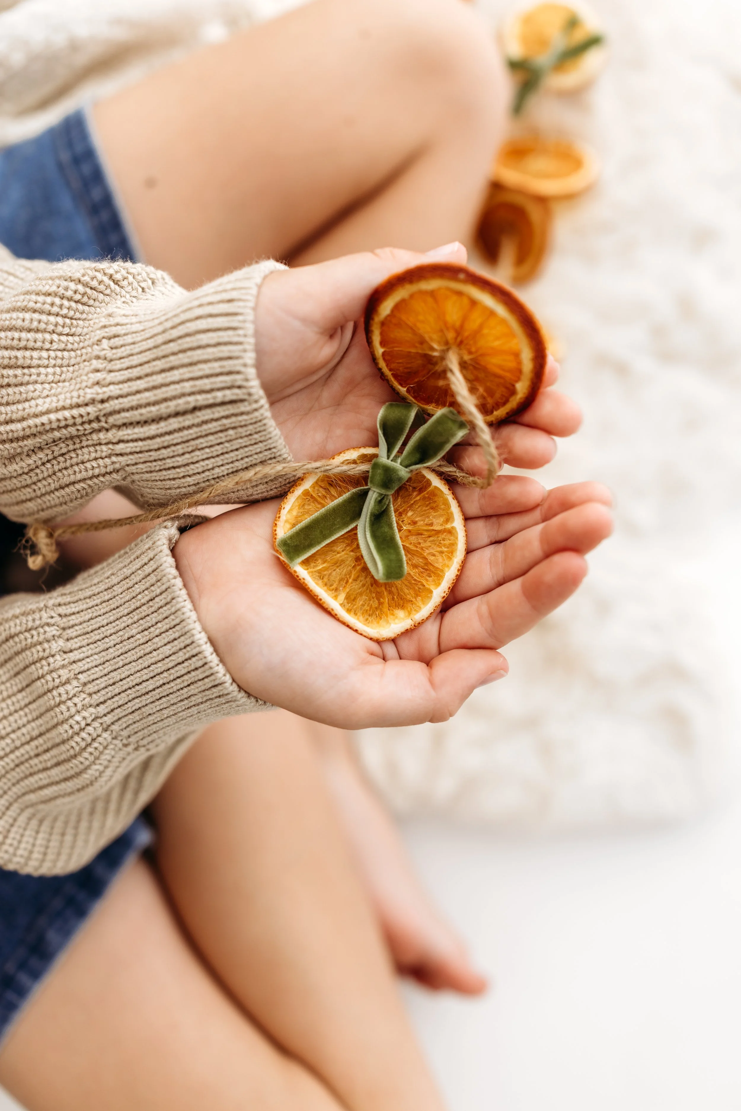 Person holding dried orange slices with green velvet ribbon, sitting on a sandy surface.