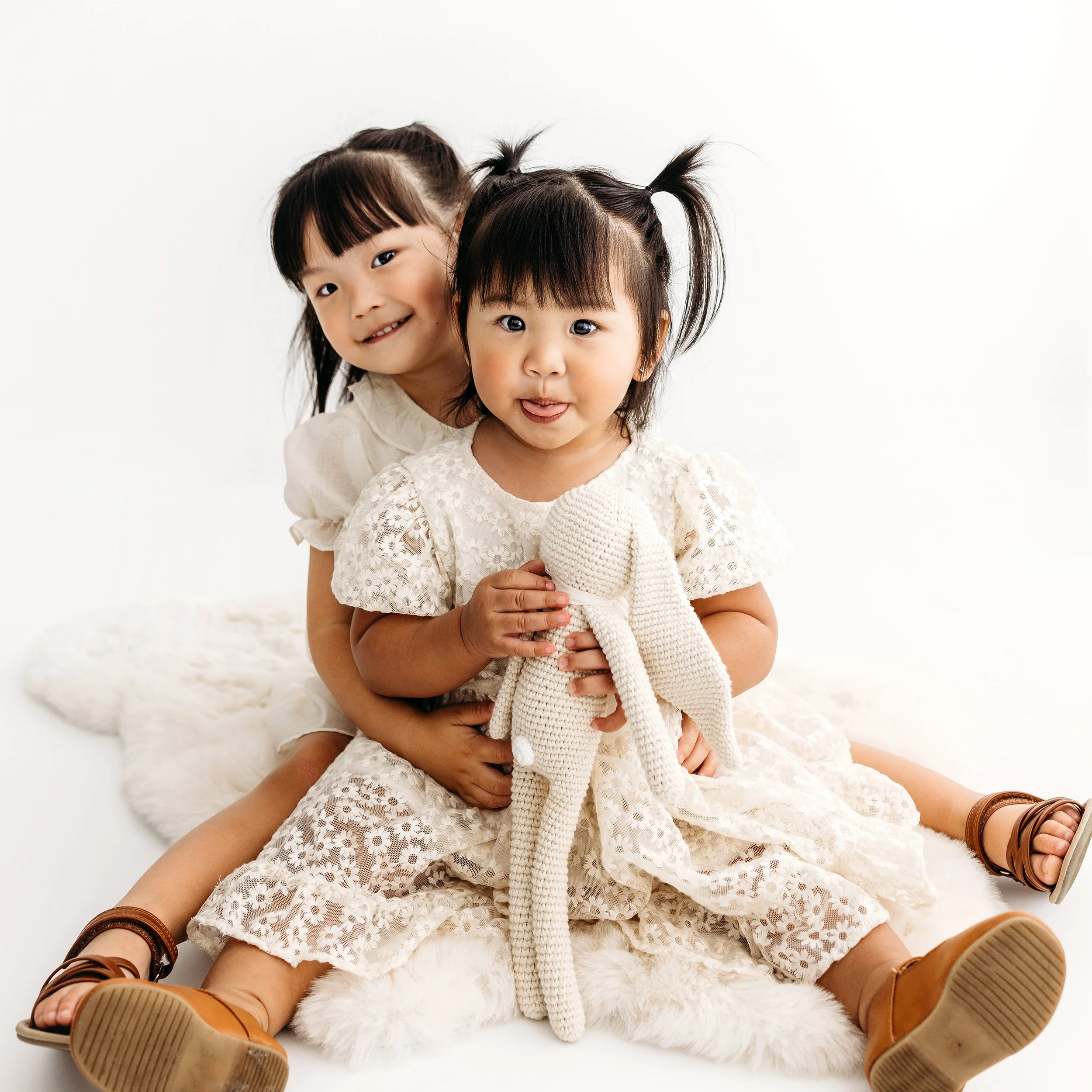 Two young sisters cuddling together in white dresses during a studio family session.