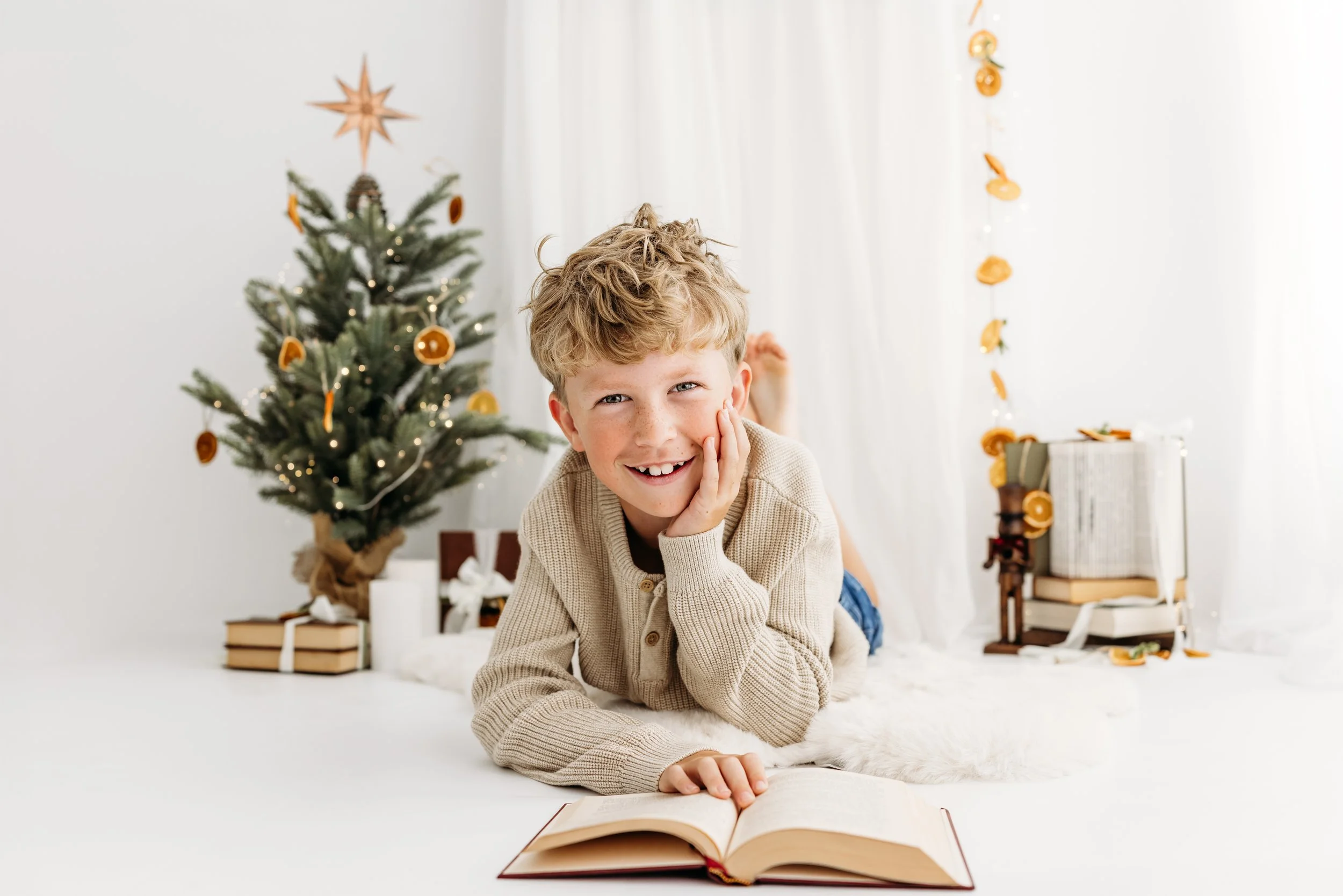Smiling boy lying with a book beside a softly decorated Christmas tree in the Aveley studio