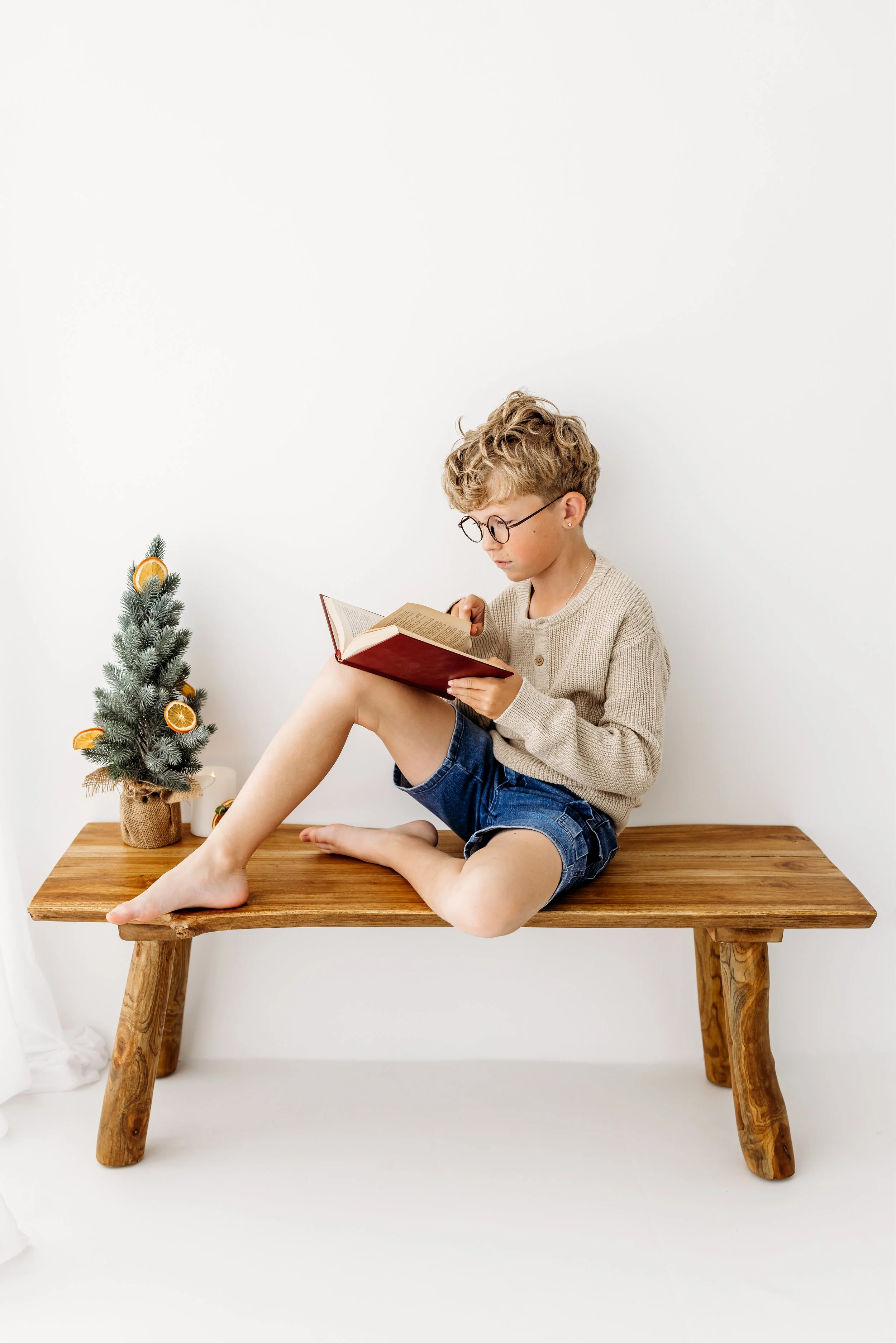 Boy reading on a wooden bench beside the Christmas tree during a relaxed studio session