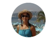 Satisfied customer of The Gleeful Ewe wearing a wide-brimmed hat and sunglasses, standing outdoors near a body of water with palm trees in the background.