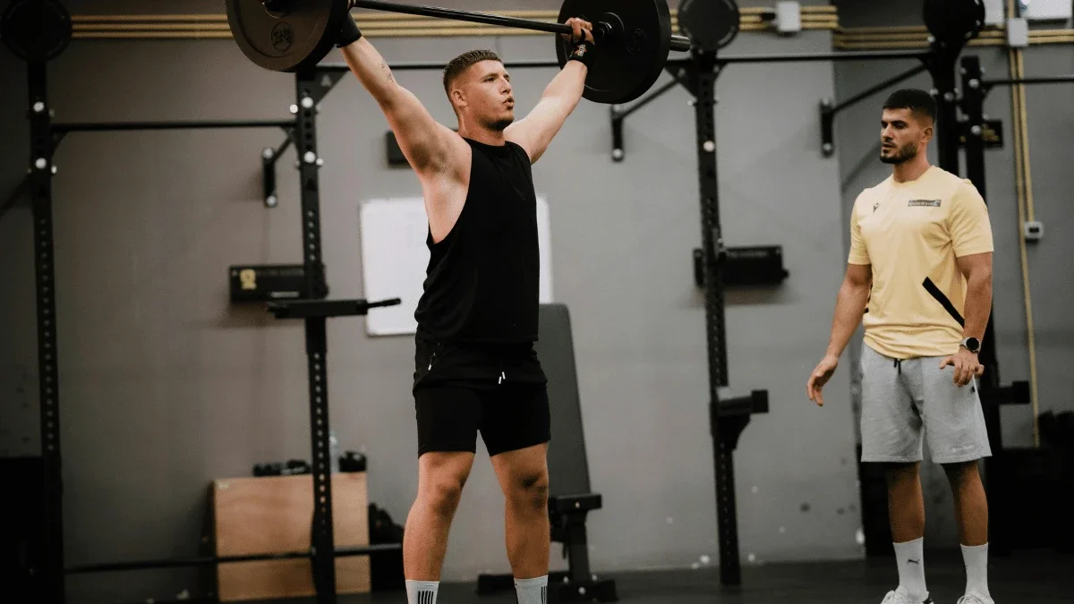 Personal trainer supervising a client performing an overhead barbell lift during a coached strength training session.
