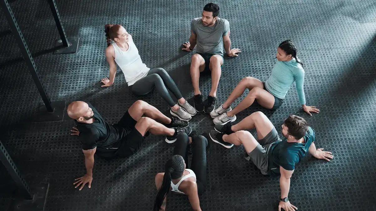 Group of adults seated in a circle on a gym floor during a fitness class discussion or mobility session, representing teamwork, accountability, and workplace wellness programs.