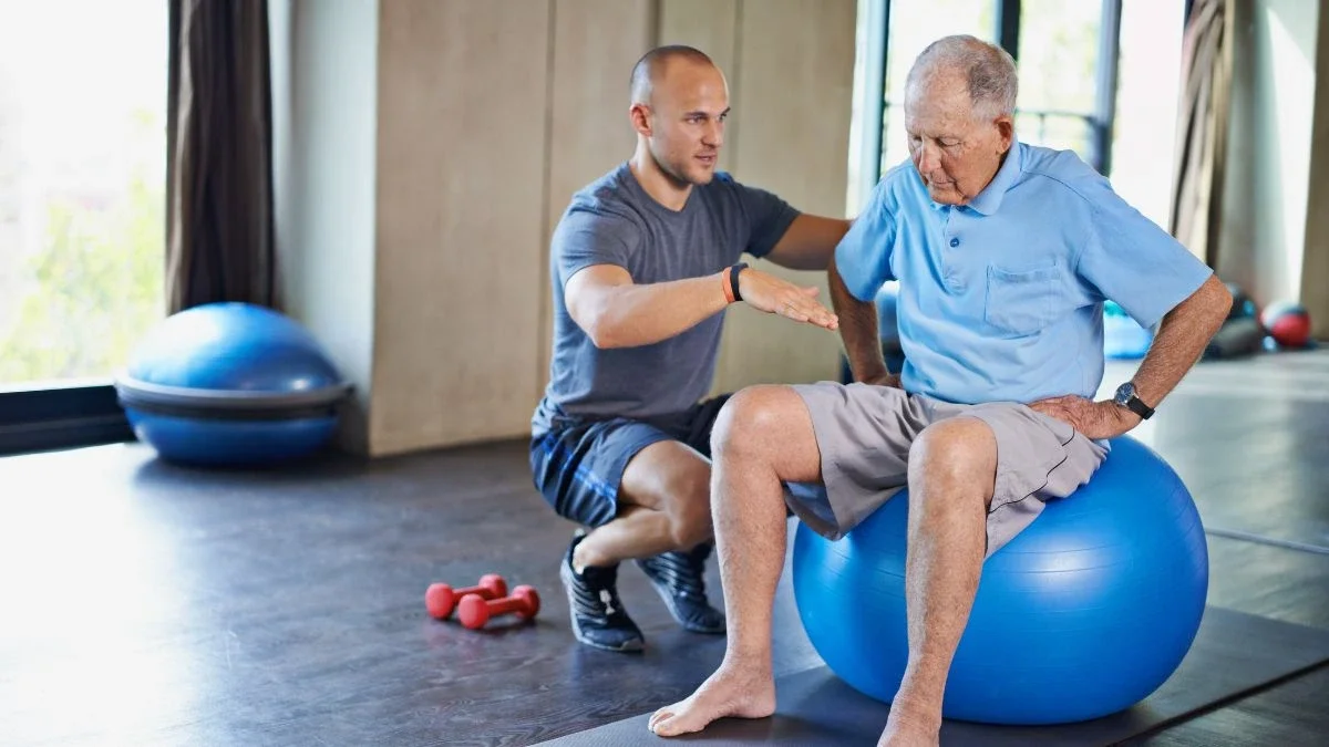 Senior sitting on exercise ball with male trainer talking to him