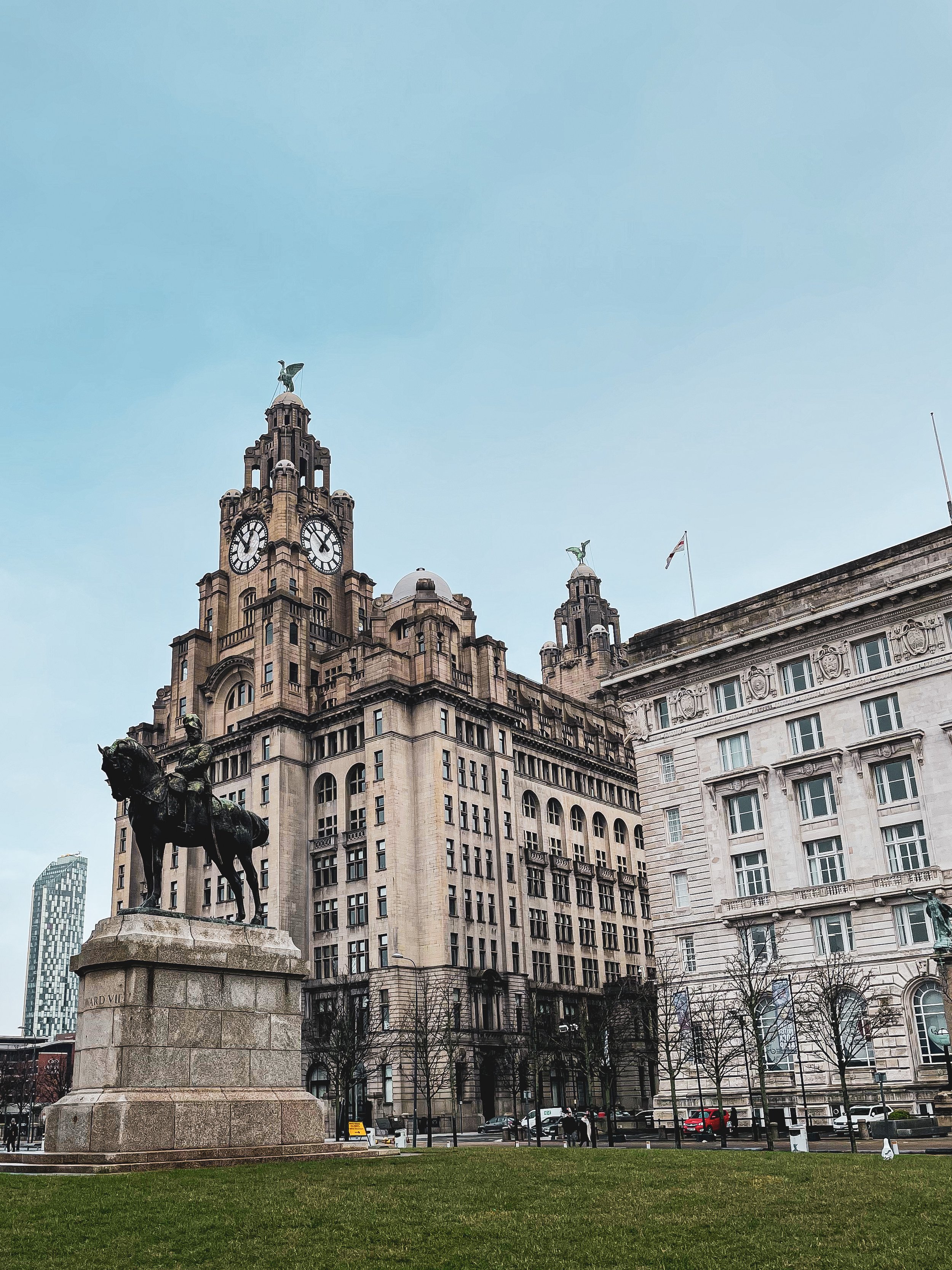 A historic building with clock towers and statues, surrounded by trees and parked cars, under a blue sky.