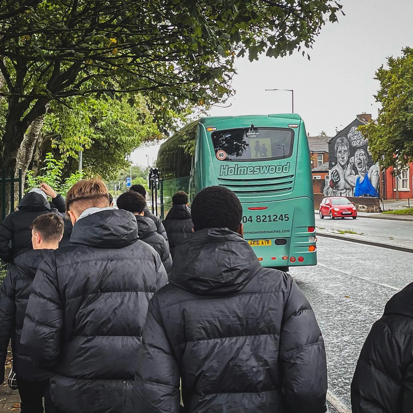 A group of people dressed in black jackets standing in line on a sidewalk next to a green bus with the word 'Homeswood' on the back. There are trees overhead and a mural with portraits on a building in the background.