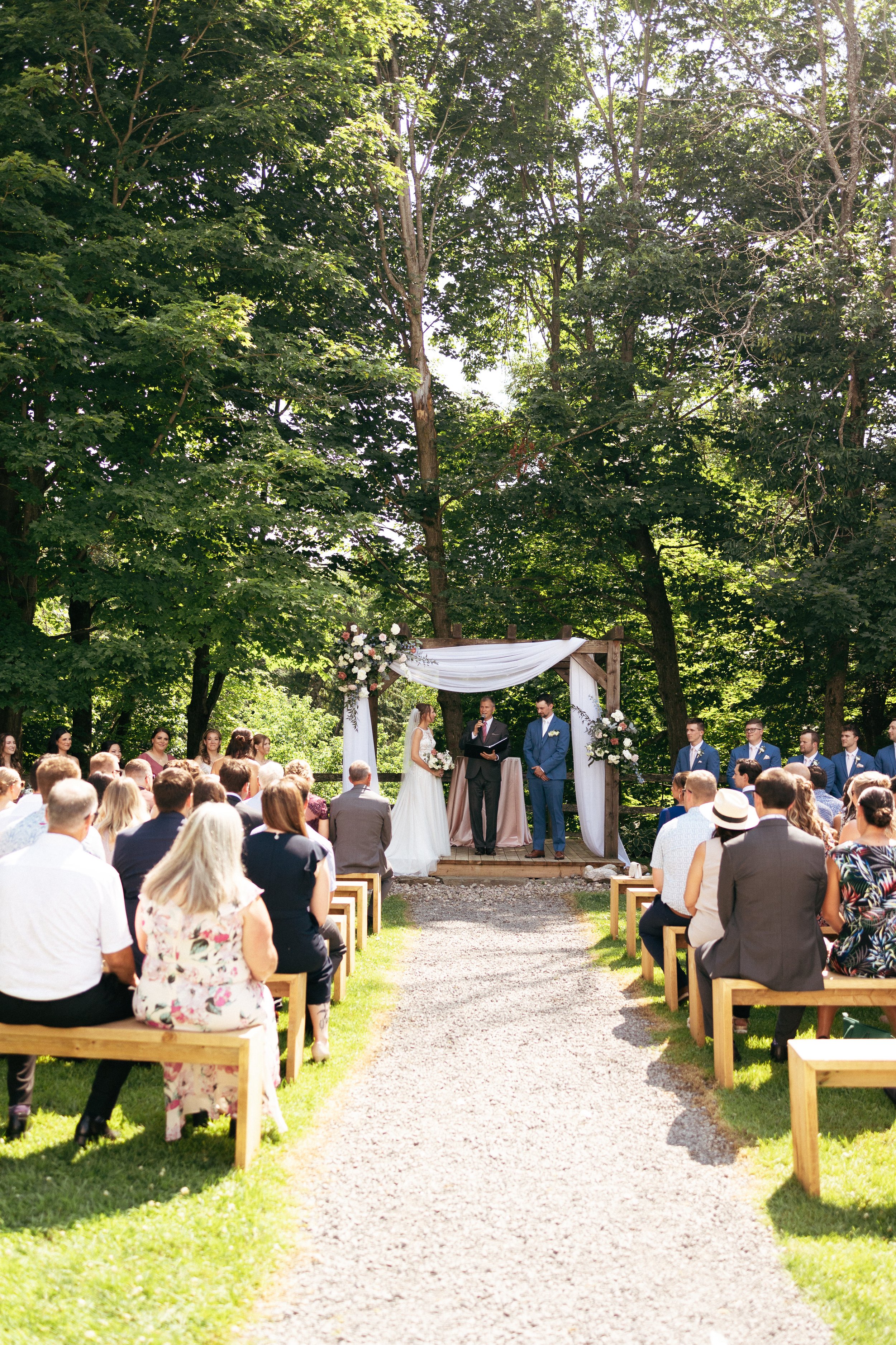 Outdoor wedding ceremony with a bride and groom standing at the altar, officiant speaking, guests seated on benches, surrounded by tall green trees and sunlight.