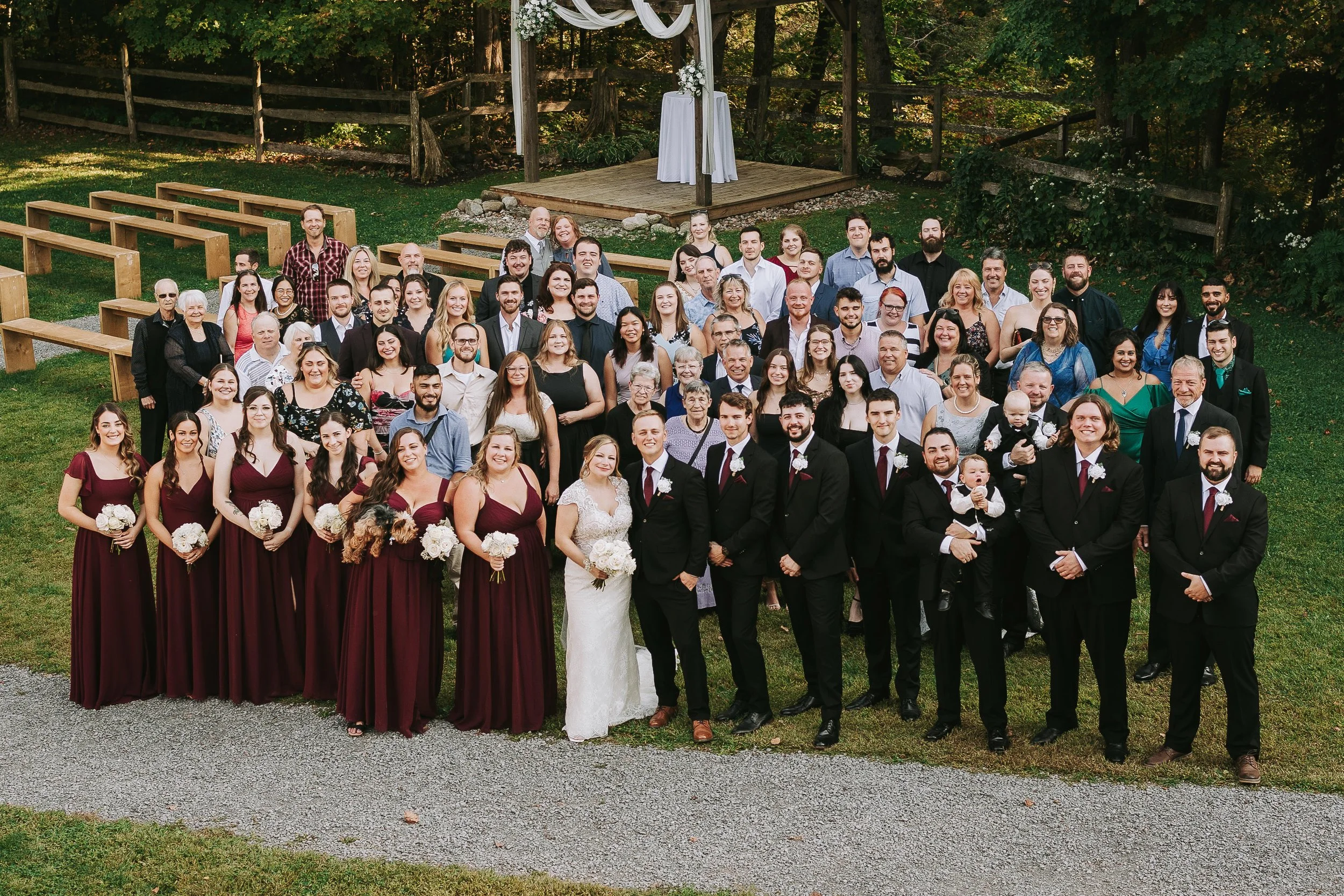 Large group of wedding guests outdoor on green grass, with a wooden stage and trees in background, all facing camera for a group photo, including bride and groom in front center, bridesmaids in burgundy dresses, groomsmen in black suits, and children