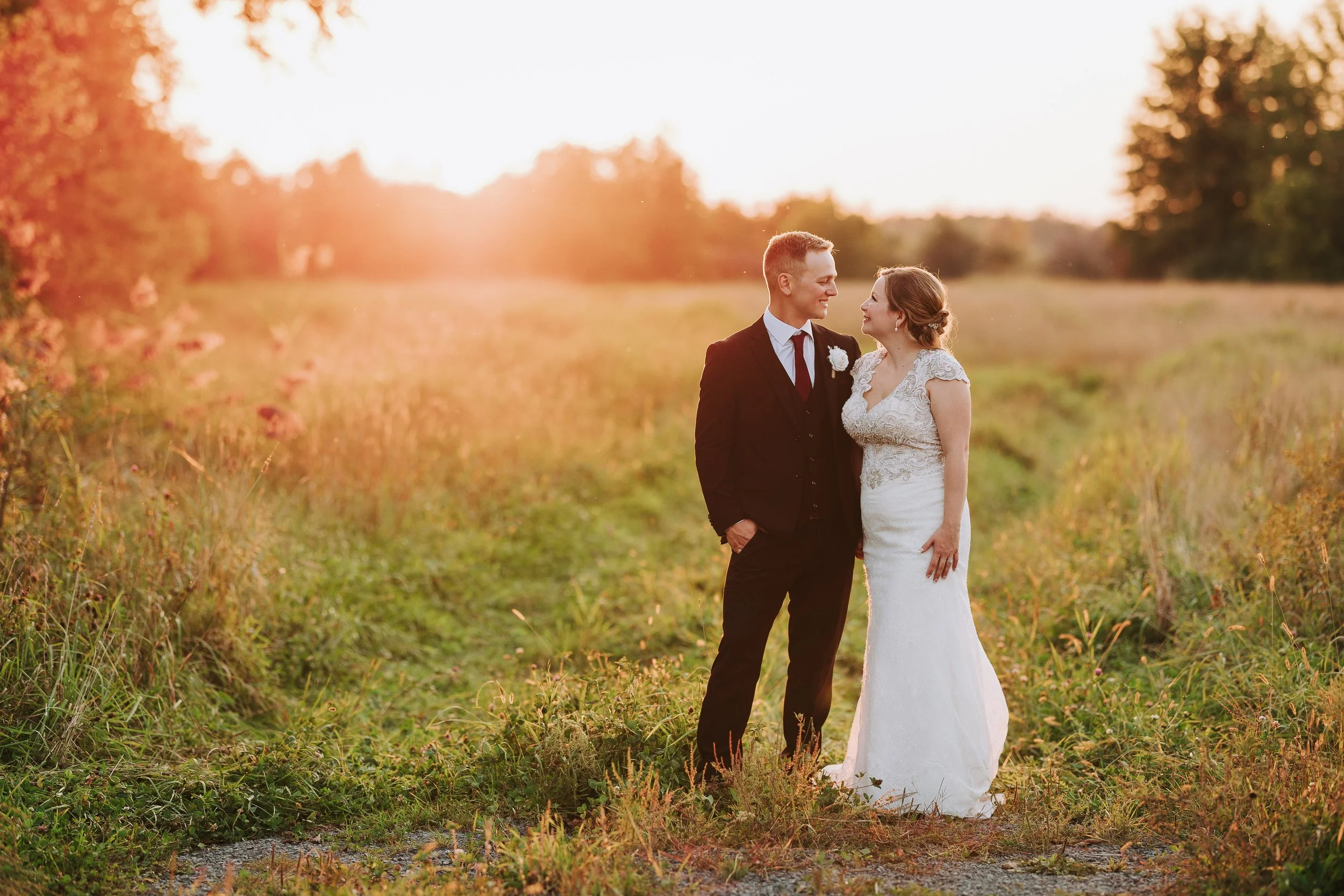 A newlywed couple stands on a grassy path in a field at sunset, gazing at each other fondly. The man wears a black suit with a white shirt and a dark tie, while the woman wears a white lace wedding dress. Trees and warm sunlight create a romantic atm
