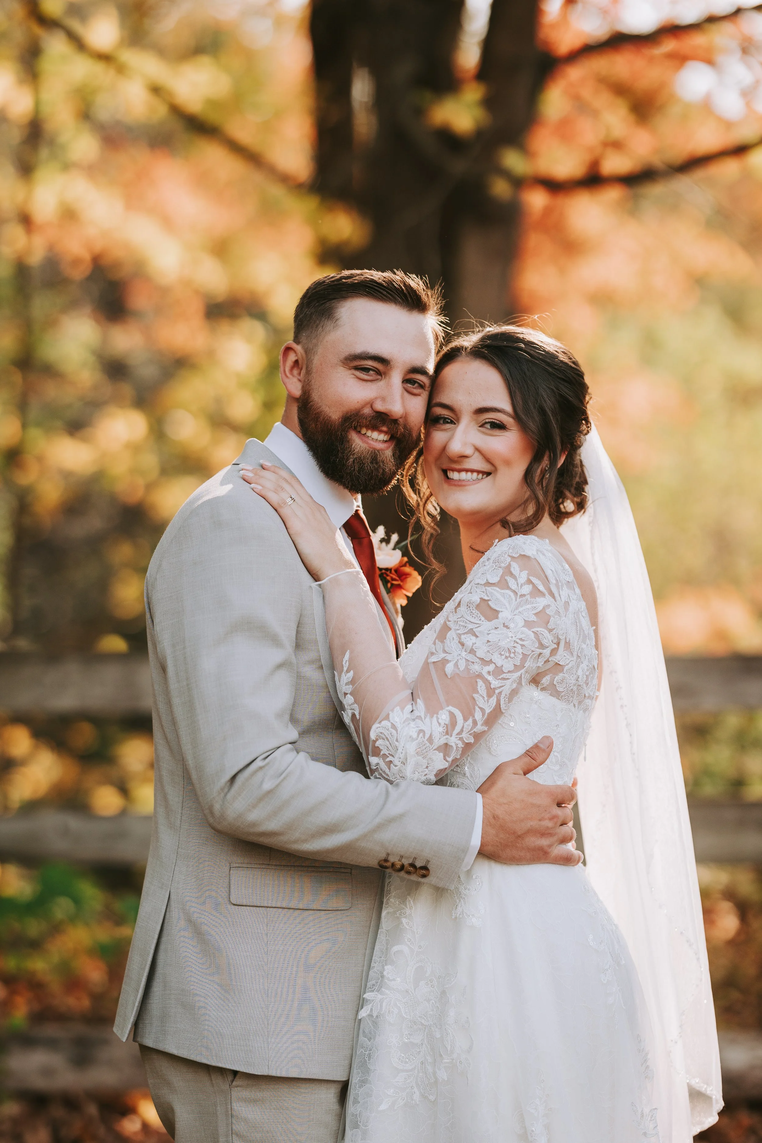 A happy couple on their wedding day, embracing outdoors in front of a tree with autumn-colored leaves, smiling at the camera.