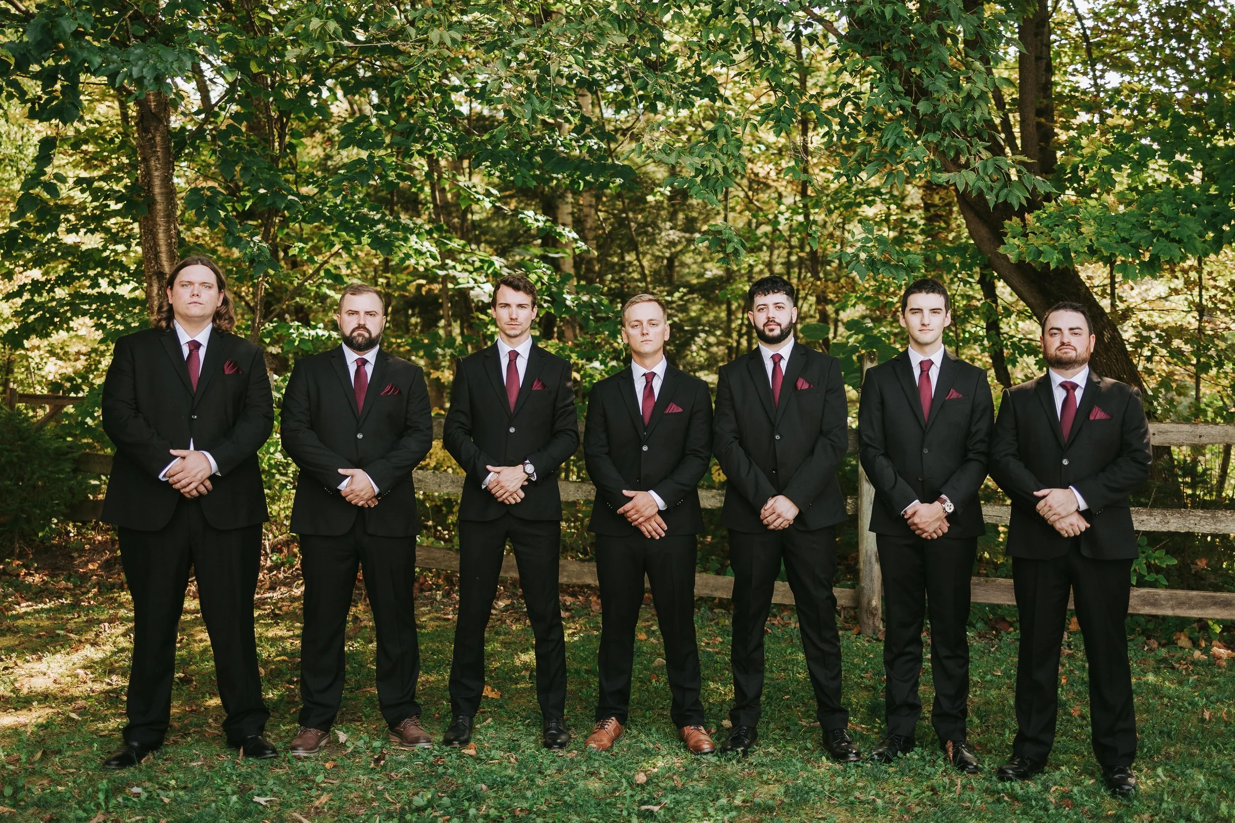 A group of seven men dressed in black suits with white shirts, red ties, and red pocket squares standing outdoors in front of a wooden fence and green trees.