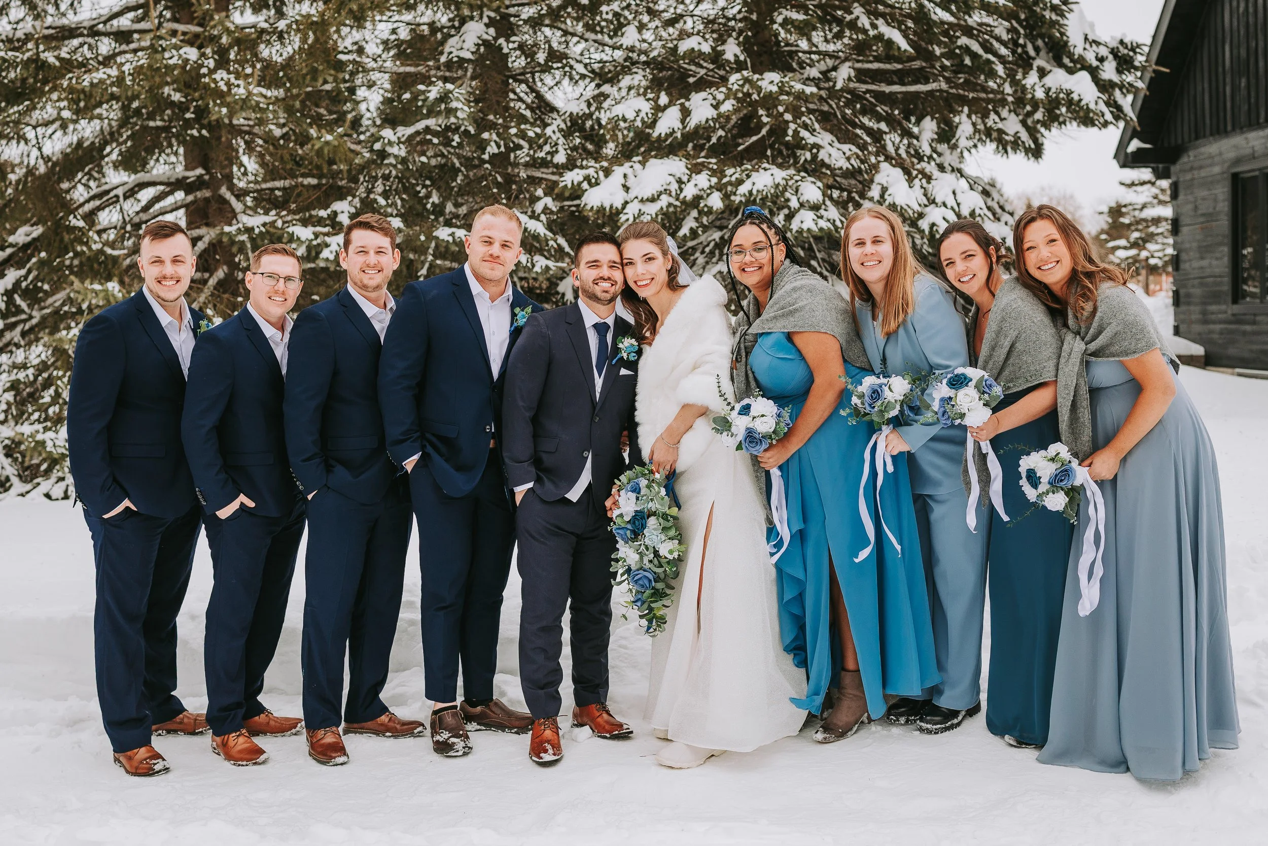A group of people dressed in wedding attire stands outdoors in snowy surroundings. The bride and groom are at the center, surrounded by bridesmaids and groomsmen, all smiling.