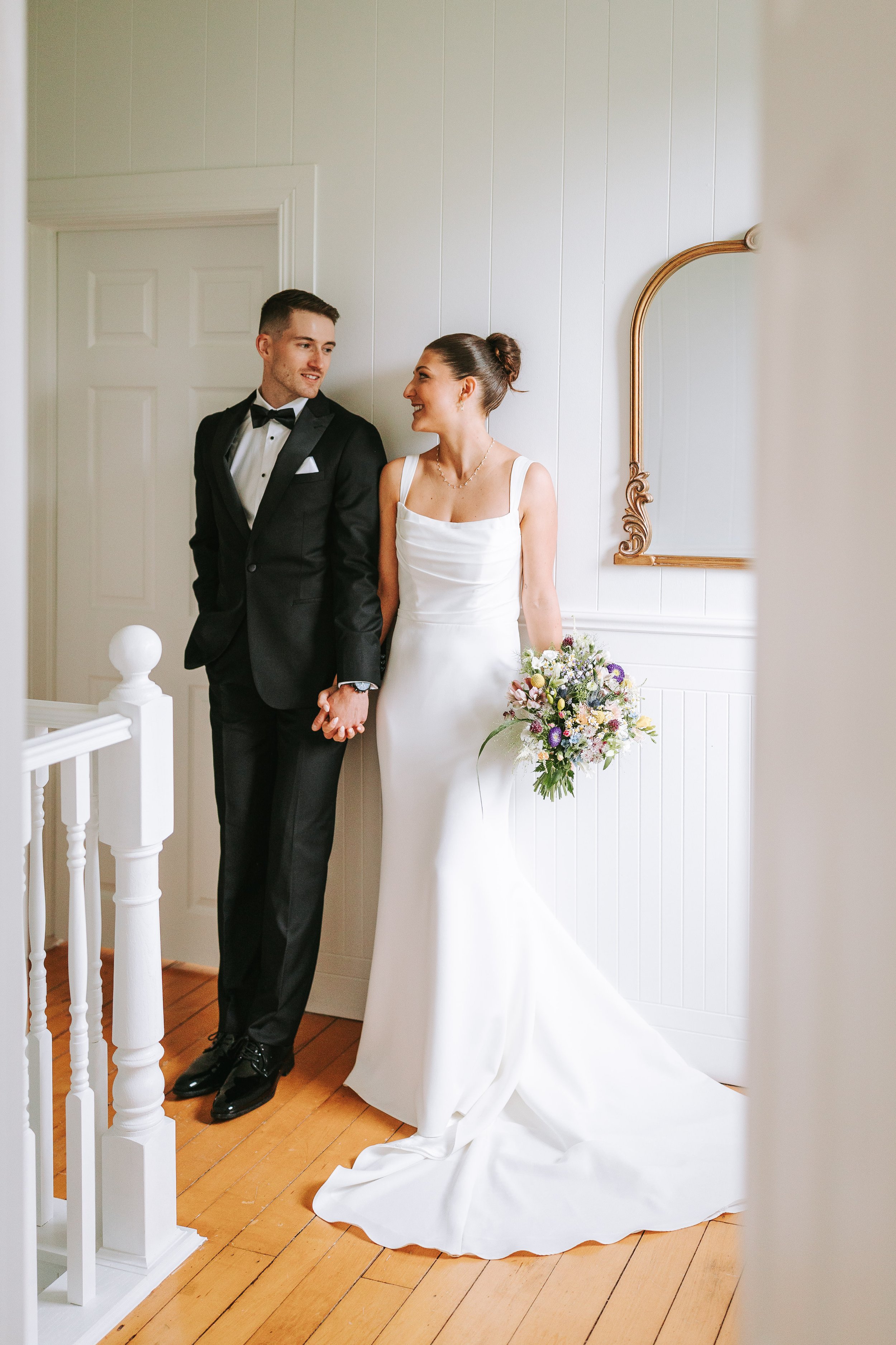 A bride and groom standing together, holding hands, and smiling at each other. The bride is in a white wedding gown holding a bouquet, and the groom is in a black tuxedo with a bow tie.