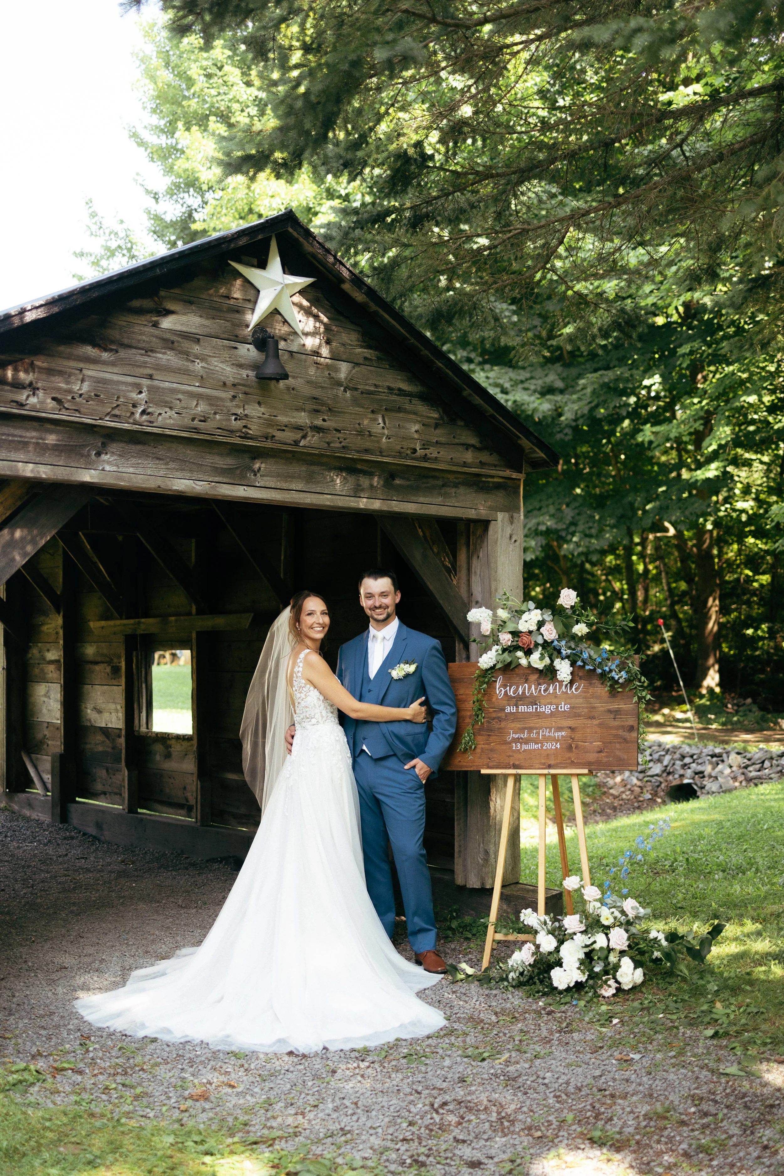 A bride and groom standing together in front of a rustic wooden structure decorated with a floral arrangement and a sign that reads 'Bienvenue au mariage de Janice et Philippe, 13 juillet 2024'. The bride is wearing a white wedding gown with lace det