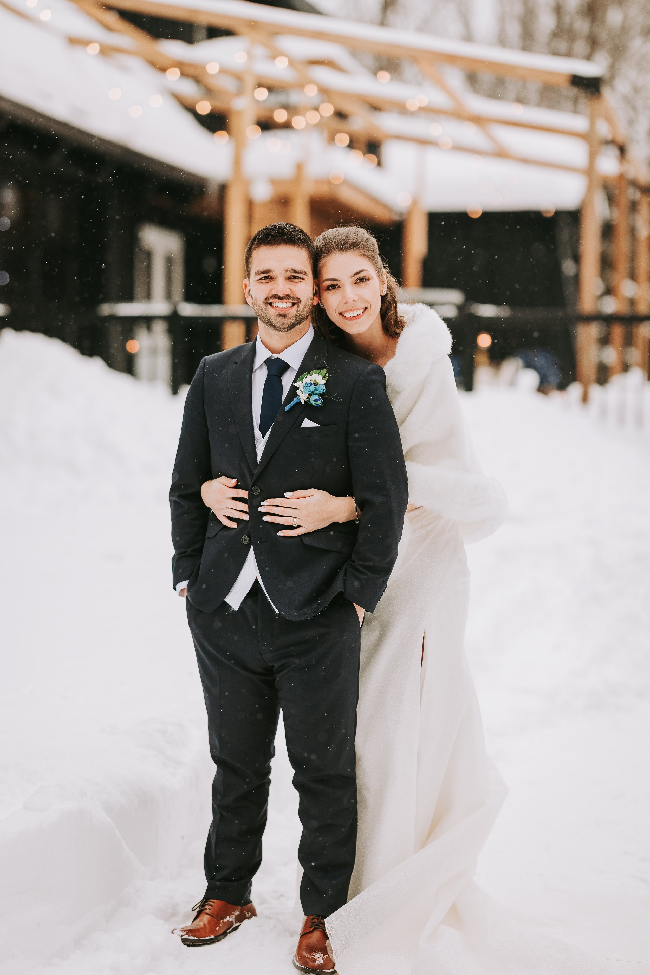 A smiling couple in wedding attire standing in the snow outside, with snow-covered ground and a building with string lights in the background.