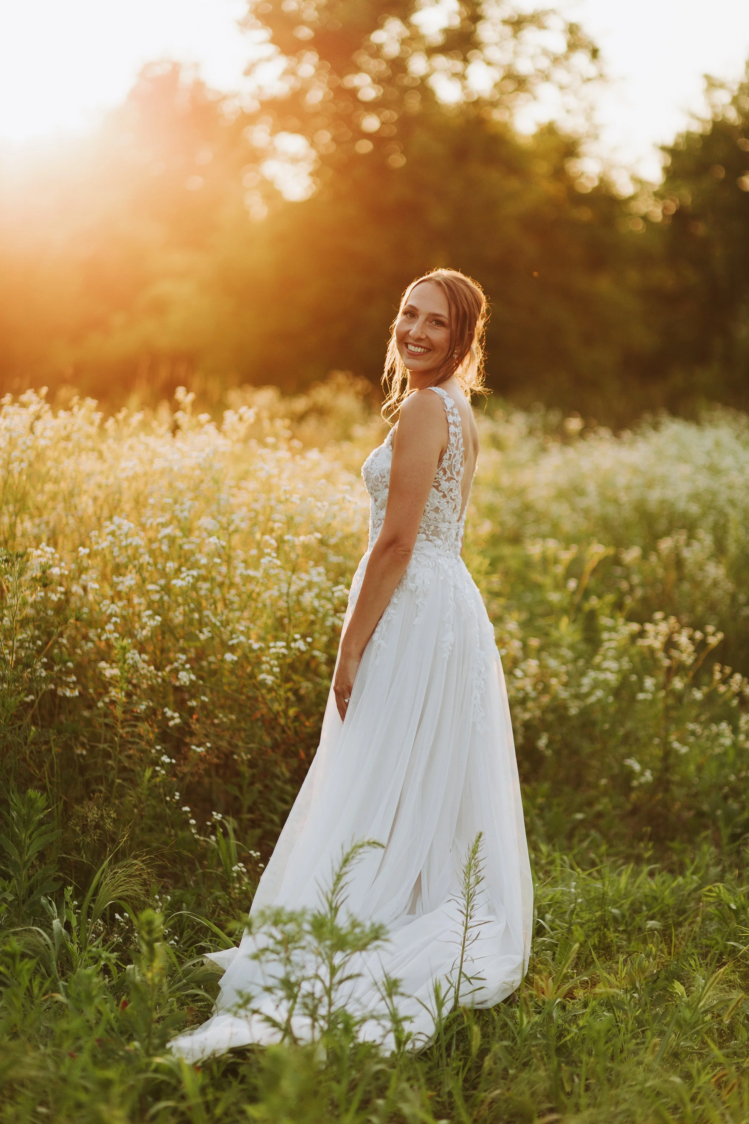 A woman in a white lace wedding dress smiling in a field of wildflowers at sunset.