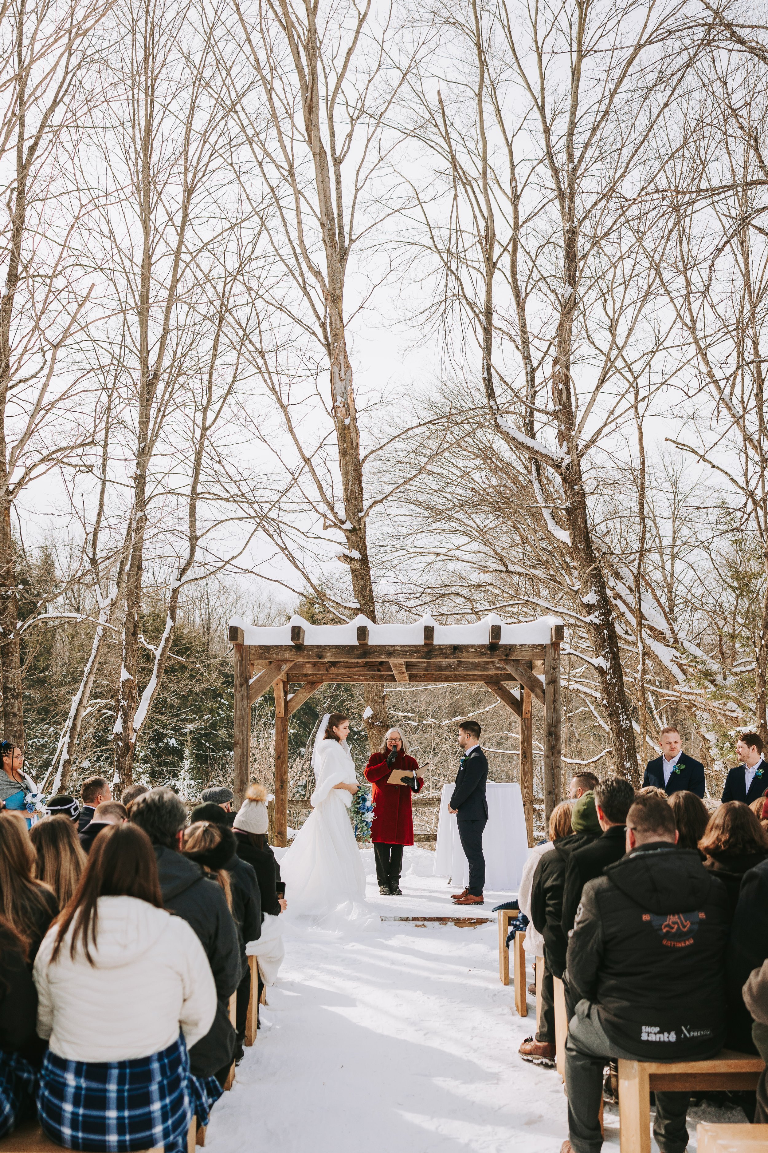 A winter outdoor wedding ceremony taking place in the snow, with the bride and groom standing under a wooden arch, surrounded by guests seated on benches, in a forested area with bare trees.