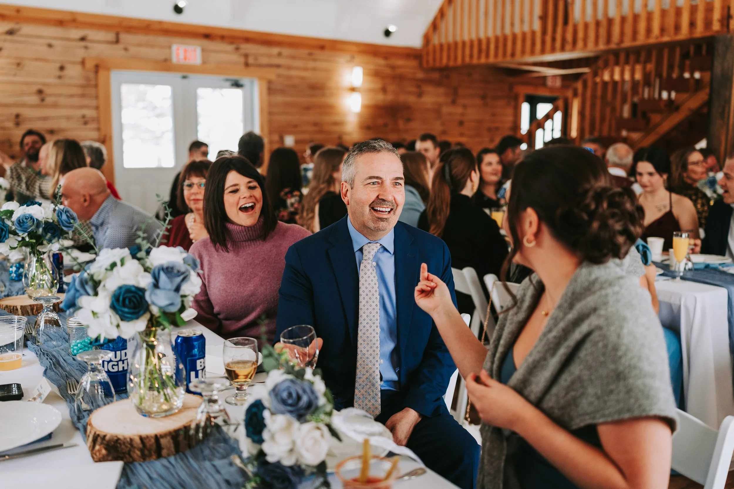 People sitting at a decorated table in a wooden banquet hall, smiling and talking, with floral centerpieces, drinks, and party supplies visible.